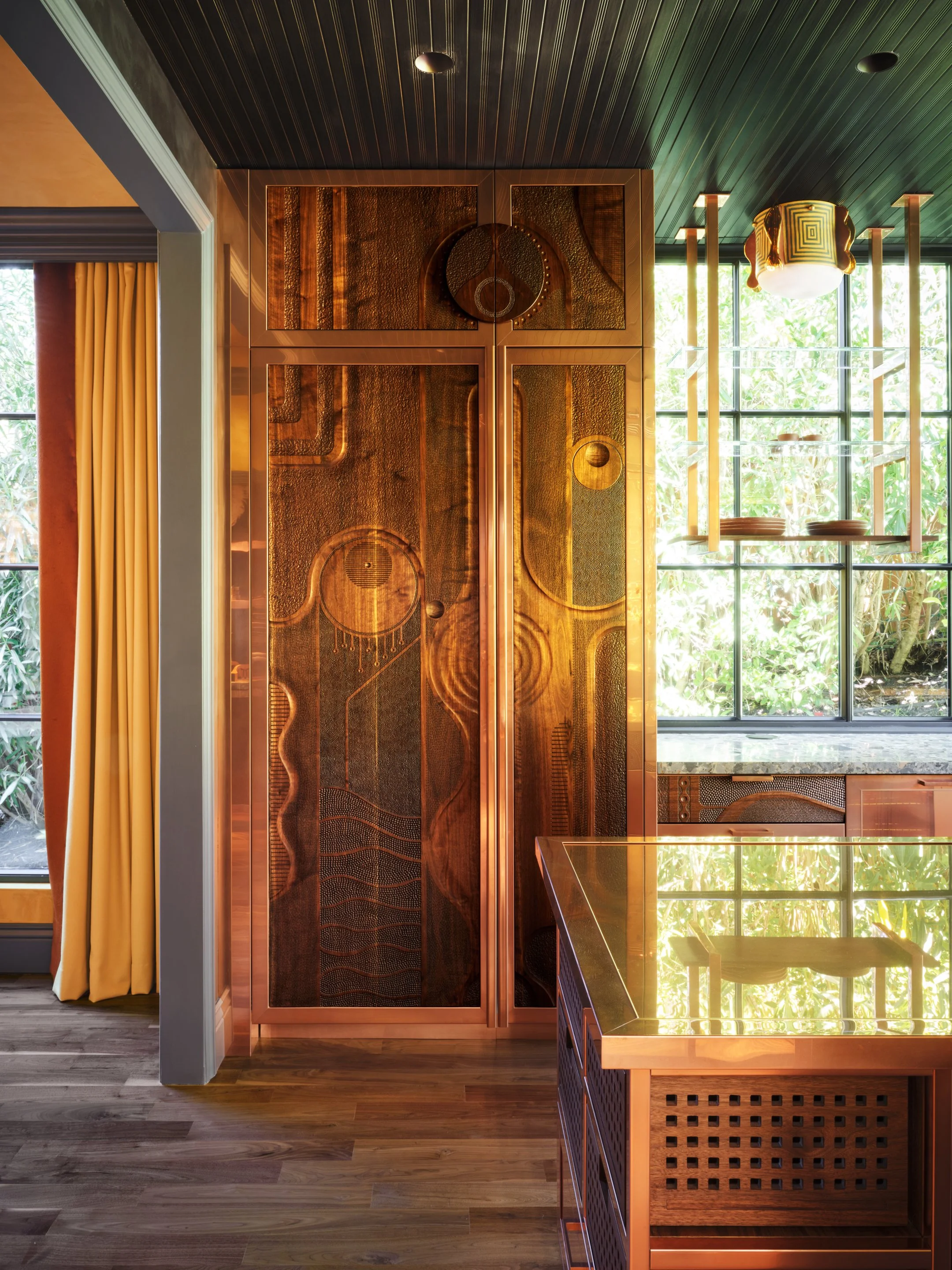 Wood and copper kitchen island and cabinetry with carved decorative wooden panels set in a bright room with expansive grid-patterned windows framing exterior greenery.