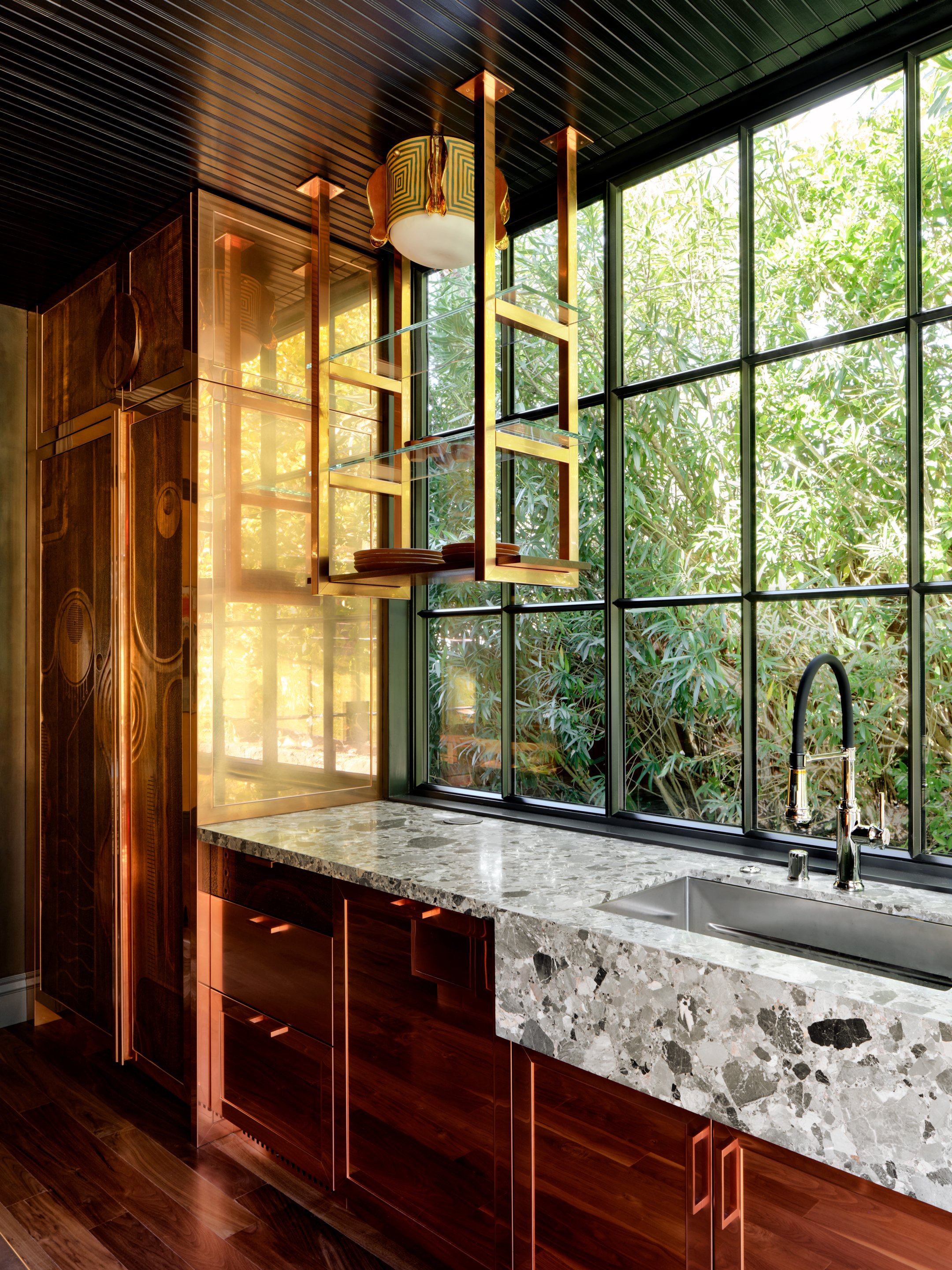 Custom kitchen millwork featuring carved wooden panels framed in copper, with large windows looking out to greenery.