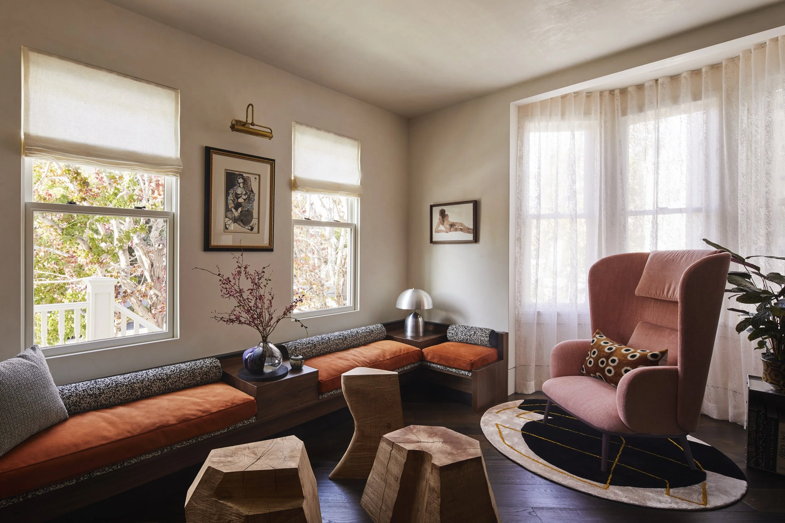 Seating area with three small, irregular wooden tables, a pink armchair, and built-in seating with orange cushions positioned in front of large windows with sheer drapery.