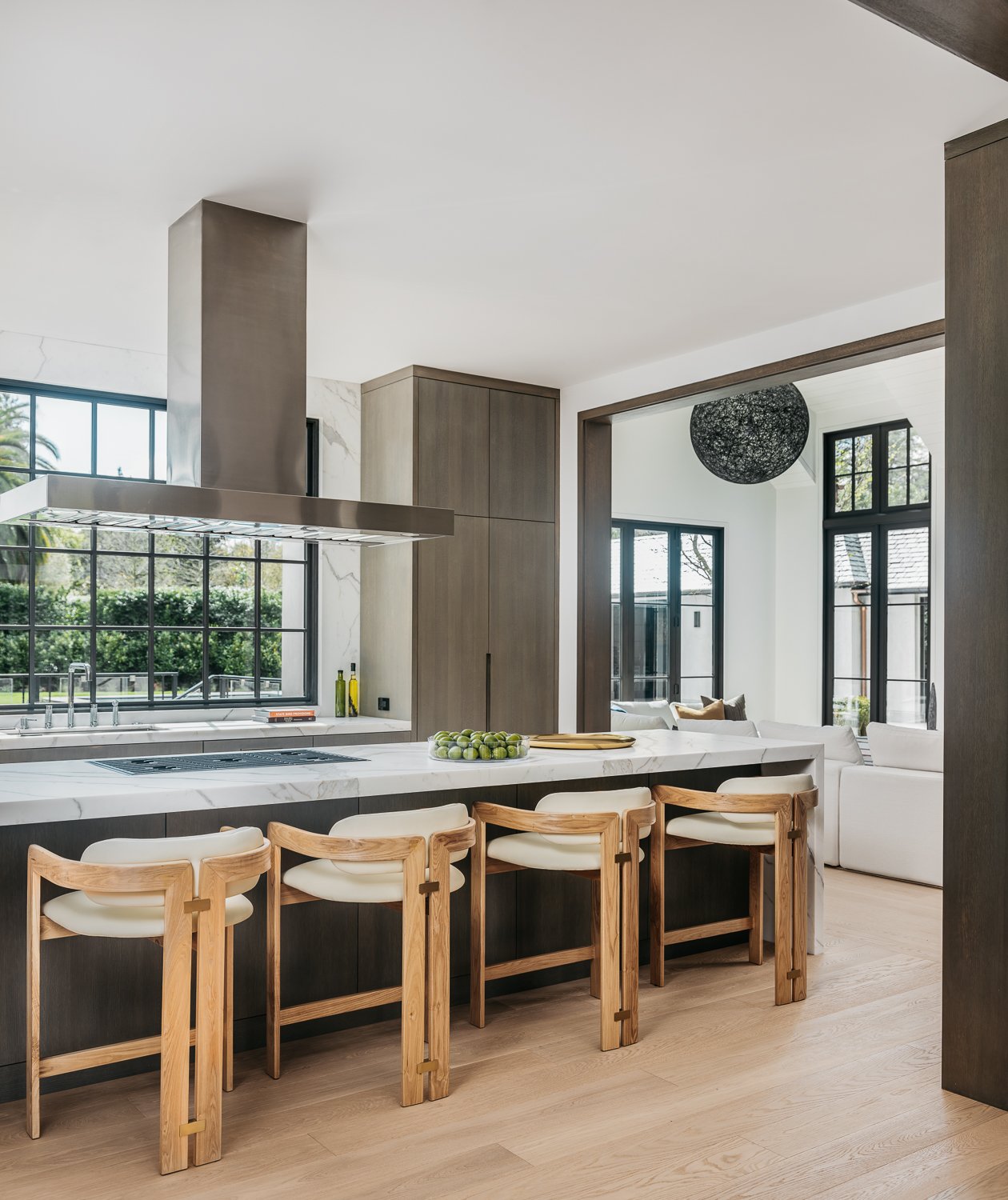 A kitchen interior featuring a central island with wood bar stools, white perimeter countertops, dark cabinetry, and large gridded window looking out to greenery; an opening leads to a living area.