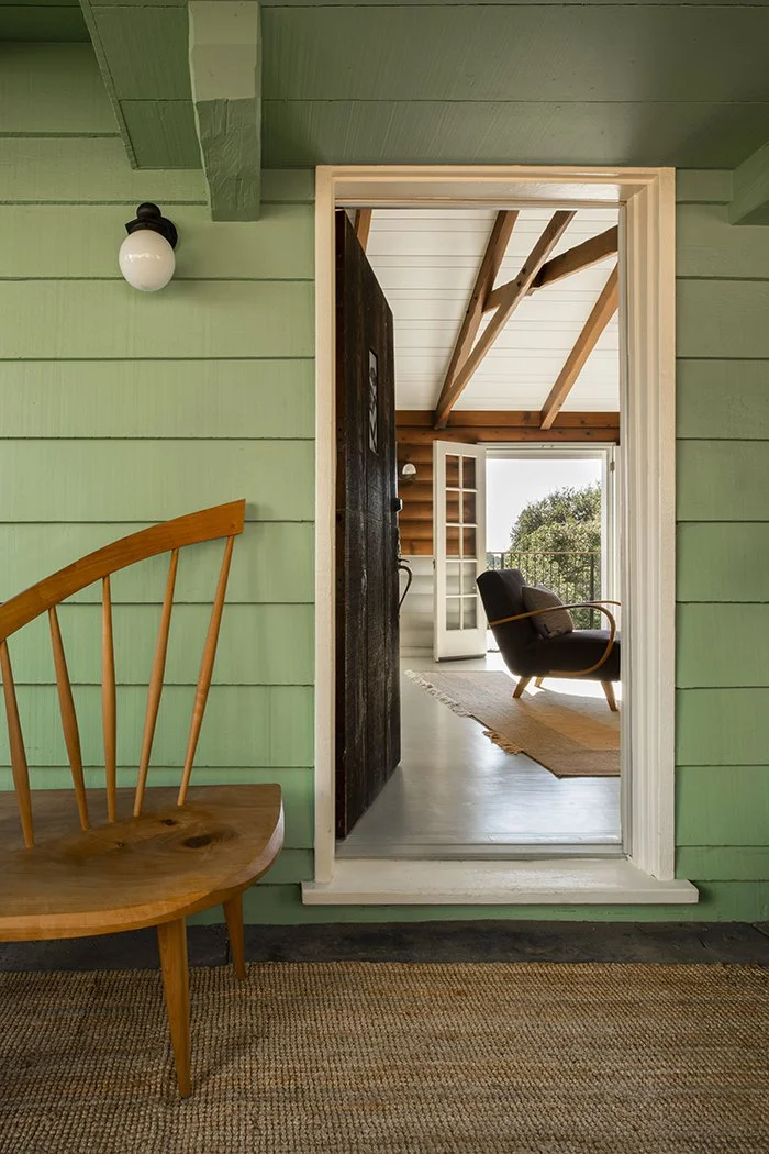 A wooden bench sits against the cabin’s green horizontal wooden siding beside an open dark wood front door; a mature oak is visible in the distance through the house.