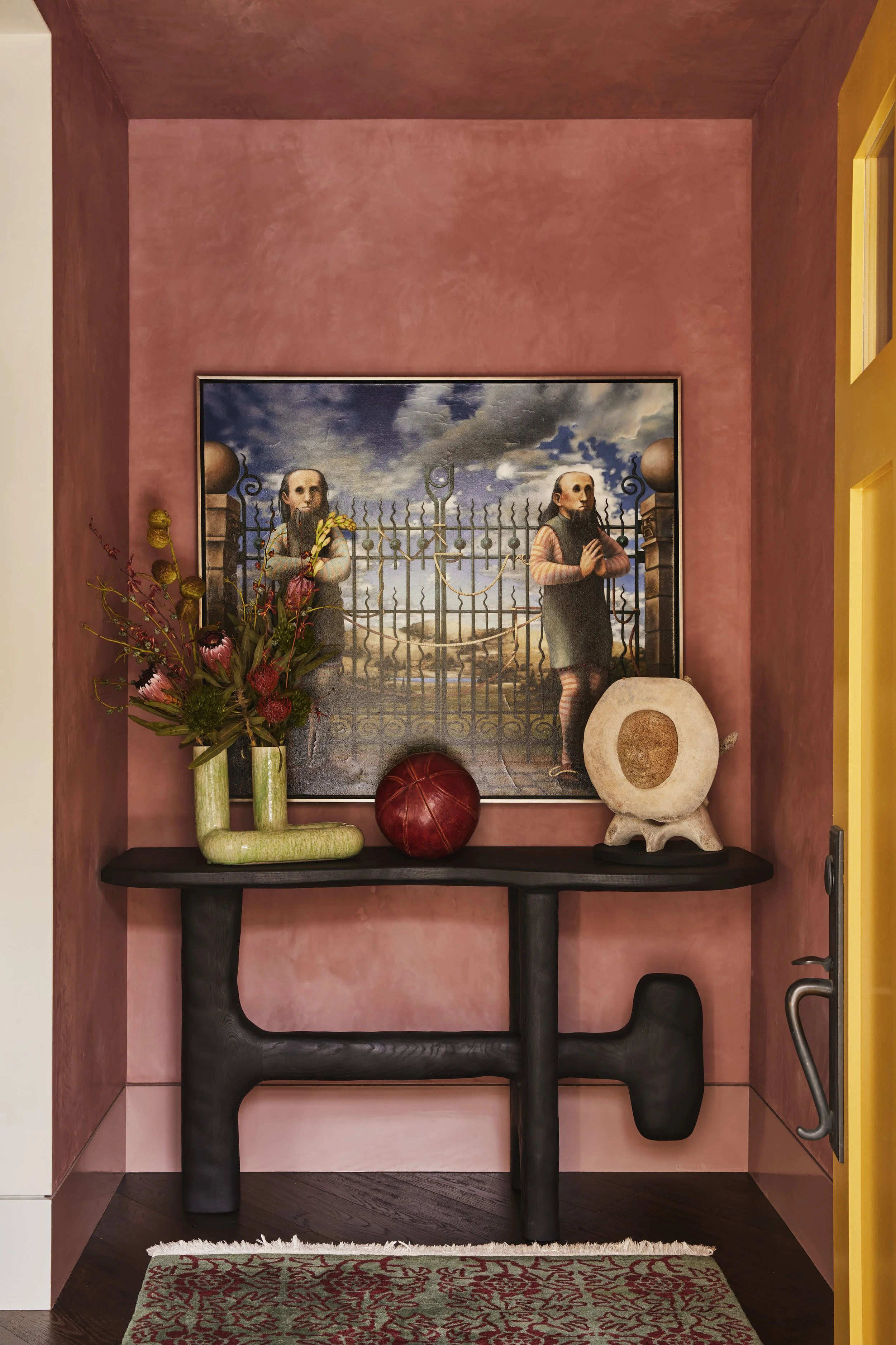 Entry vestibule featuring a sculptural wood table, red textured walls, a warm yellow front door, and a painting.