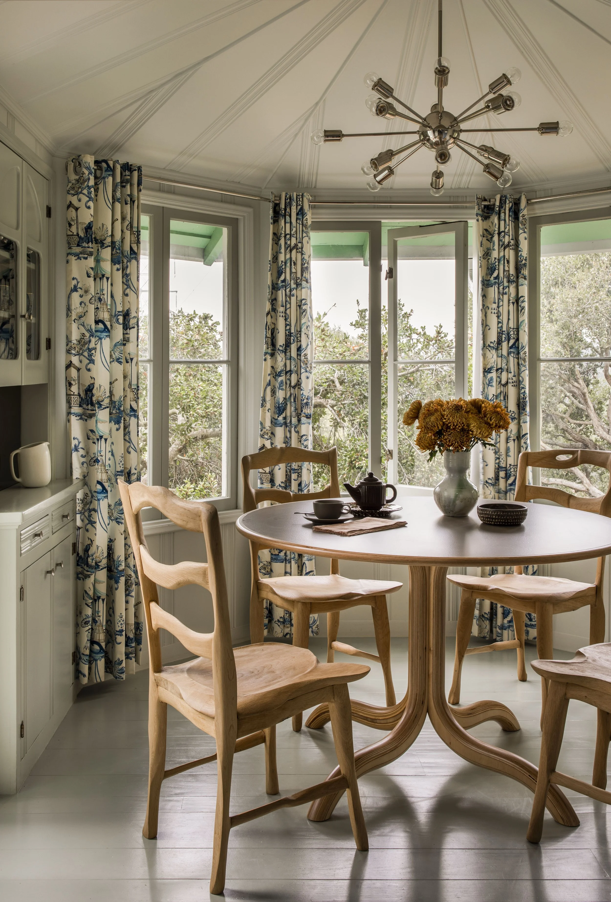 Charming breakfast nook featuring hand-carved wooden dining chairs by French furniture maker Paul Salet surrounding a wooden table, illuminated by a modern metal chandelier, with patterned curtains framing the windows looking out onto green trees.
