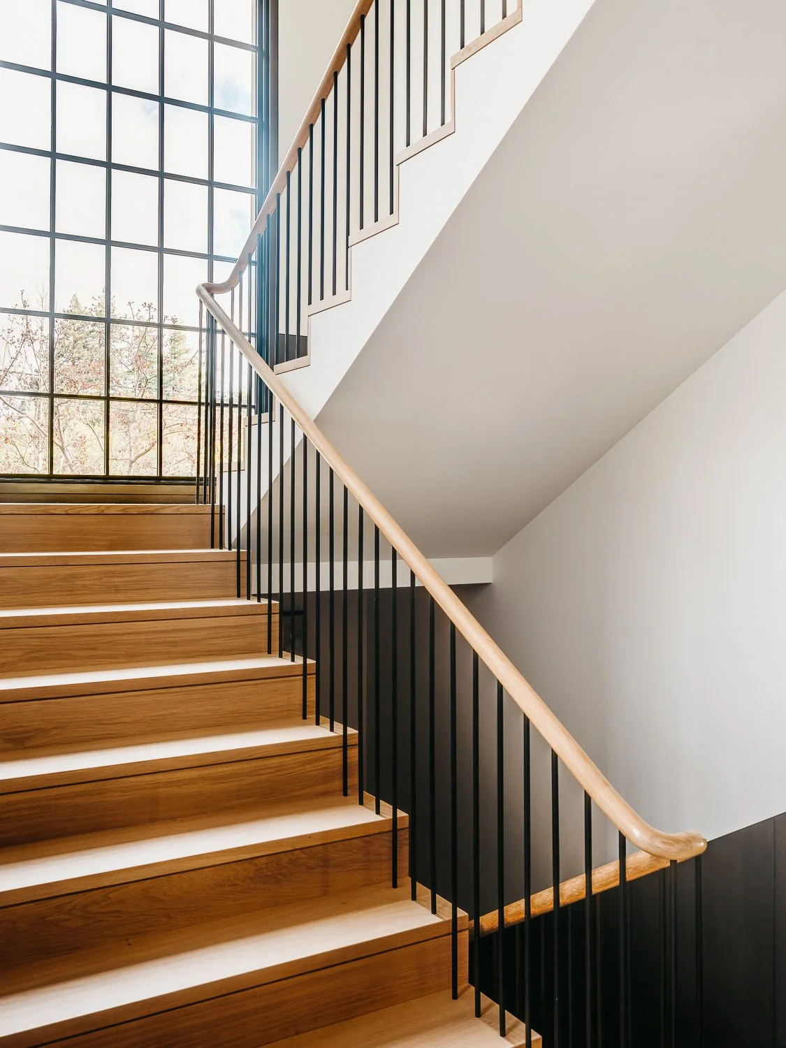 An interior staircase with wooden treads, a dark metal railing with wooden handrail, and tall vertical gridded windows with treetop views.
