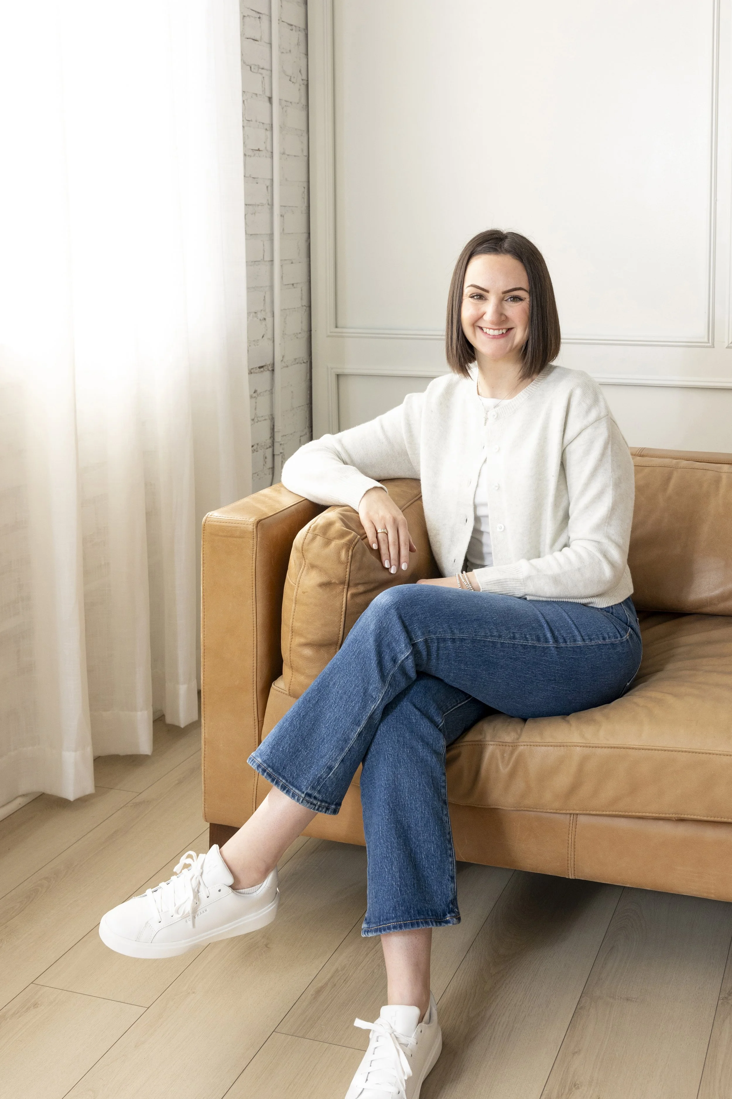 A woman with shoulder-length brown hair, wearing a white sweater, blue jeans, and white sneakers, sitting on a tan leather sofa in a room with white brick wall and white curtains, smiling at the camera.