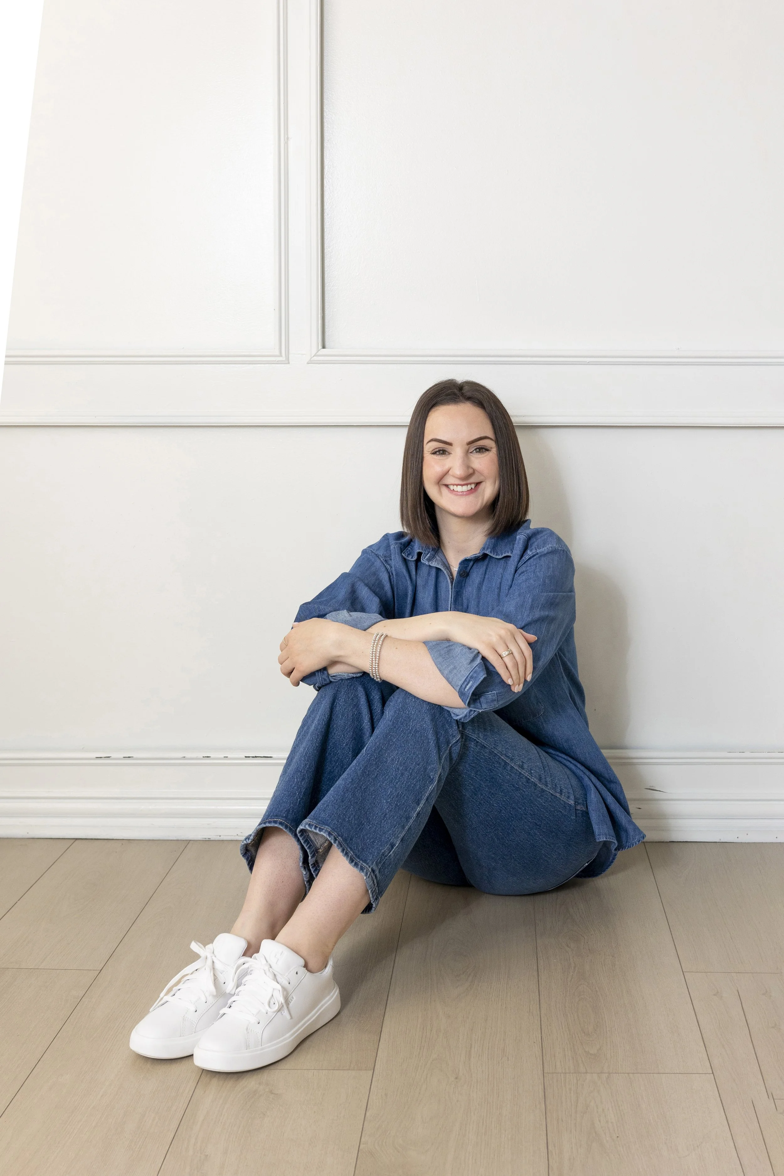 A young woman with shoulder-length brown hair, sitting on the floor against a white wall with decorative molding. She is smiling at the camera, wearing a blue denim shirt, matching jeans, white sneakers, and multiple bracelets.