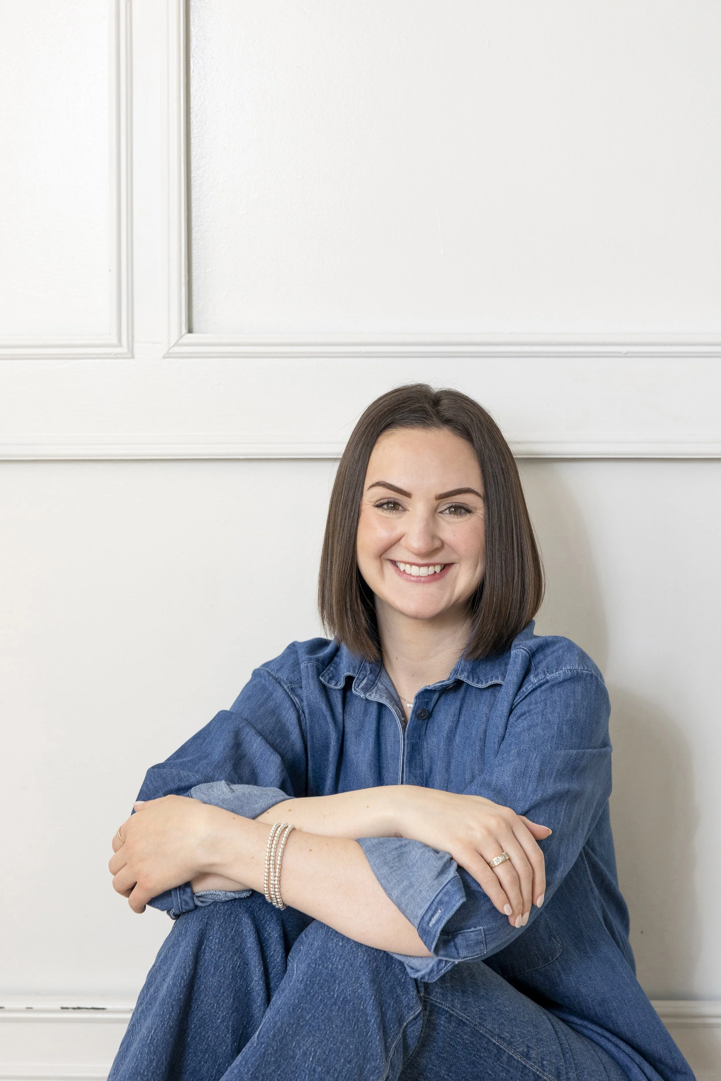 A woman with shoulder-length brown hair, smiling, sitting against a white wall, wearing a blue denim shirt and jeans.