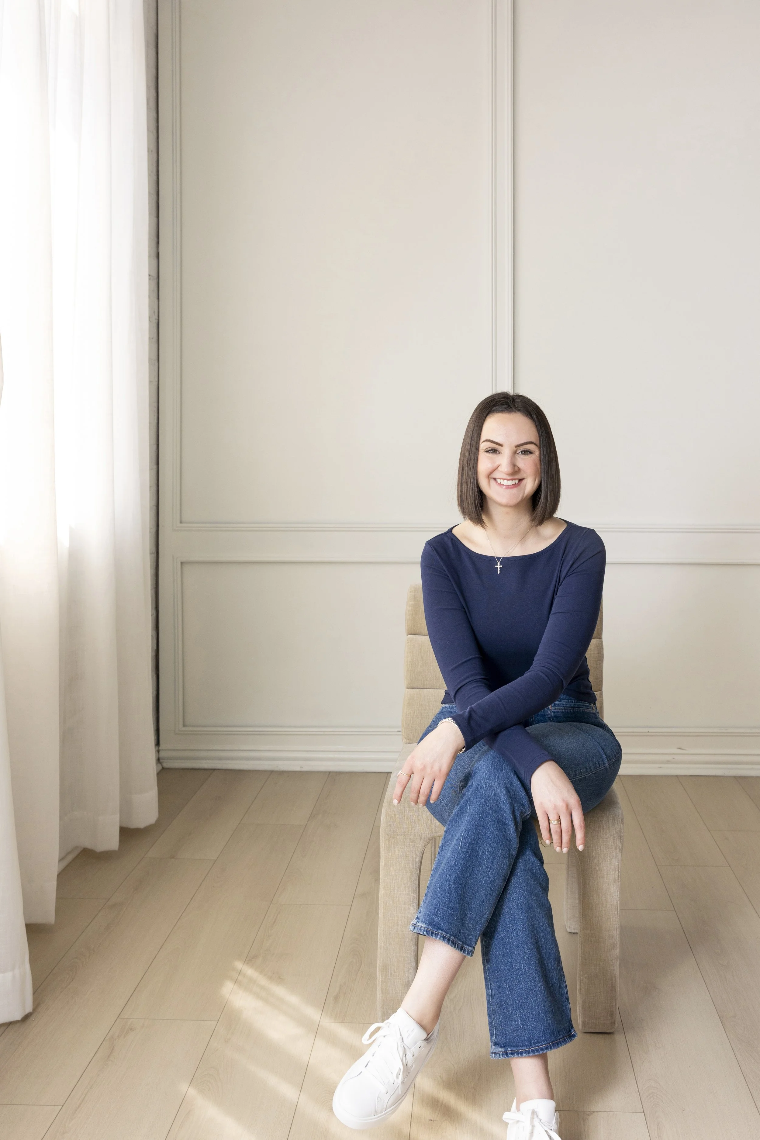 A woman with shoulder-length brown hair sitting on a beige chair in front of a light-colored wall, smiling and crossing her legs. She is wearing a navy blue long-sleeve top, blue jeans, white sneakers, and a silver cross necklace. The room has light wood flooring and a white curtain on the left side.