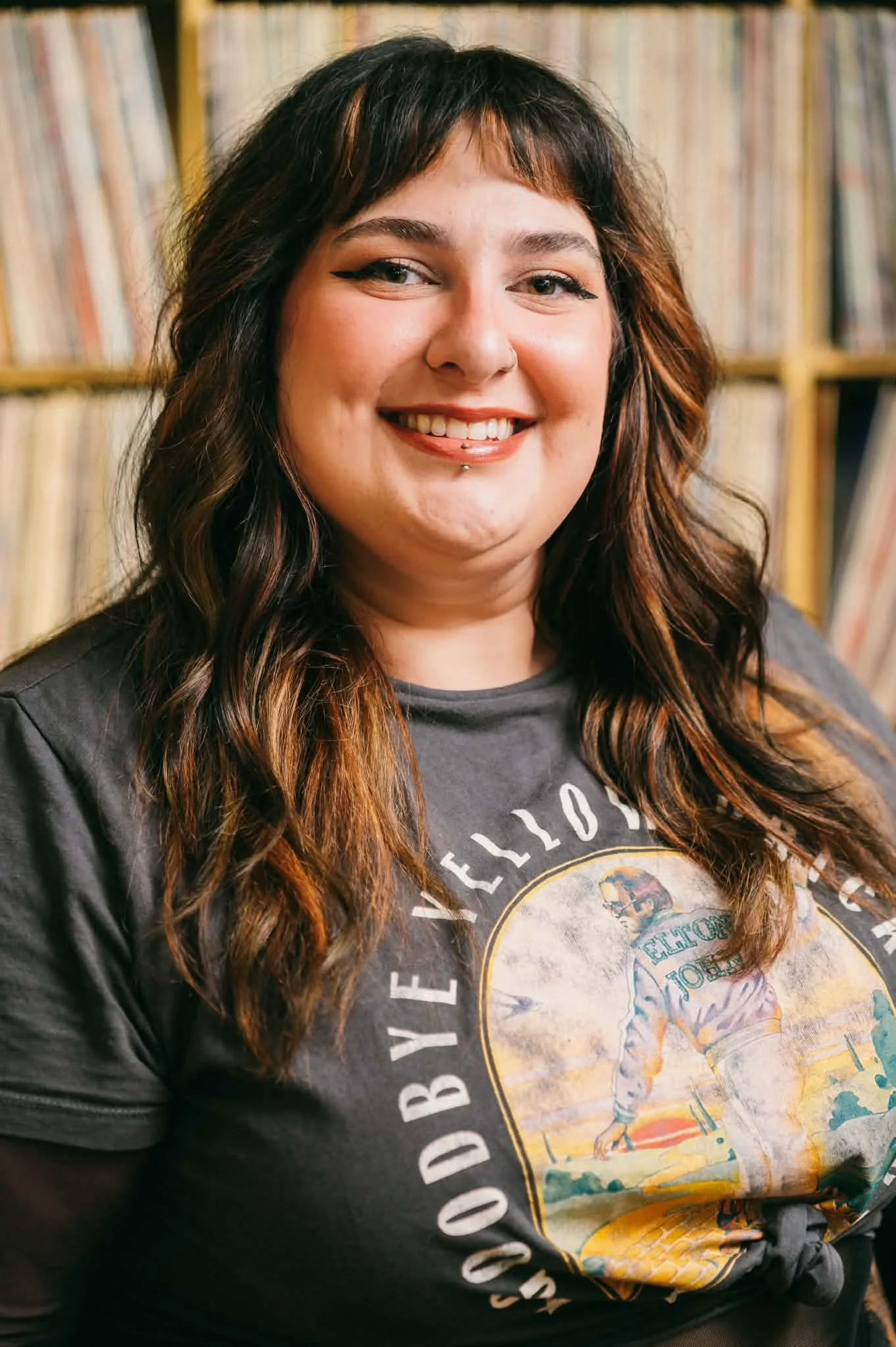 A woman with long wavy hair, dark brown with lighter highlights. She has a lip piercing and a nose ring. She is smiling and wearing a black leather jacket and a graphic t-shirt with a vintage design. She is standing in front of a yellow shelf filled with vinyl records.