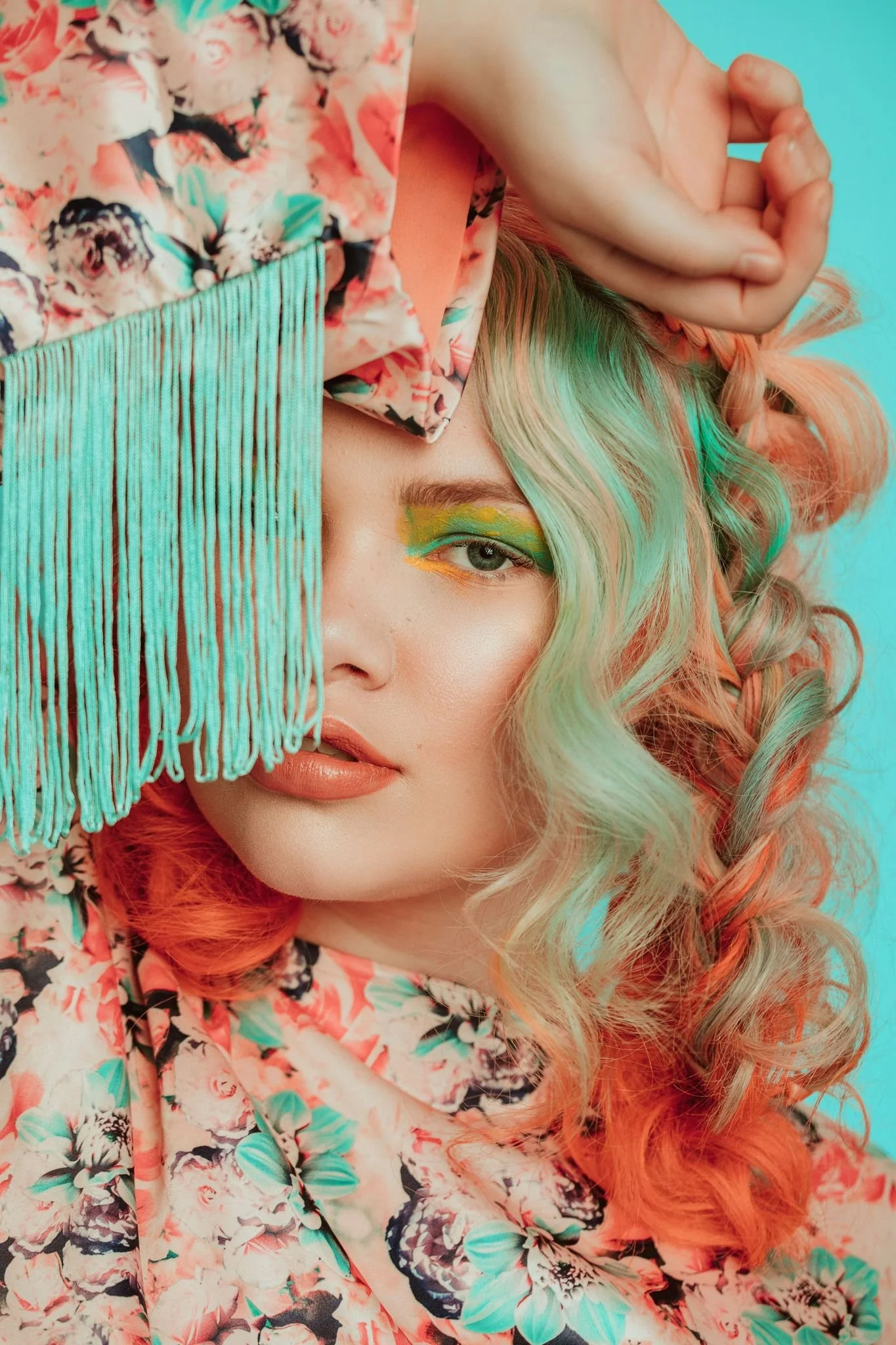 Close-up of a woman with curly, multicolored hair, wearing a floral patterned top, covering part of her face with a turquoise fringed accessory, with colorful eye makeup.