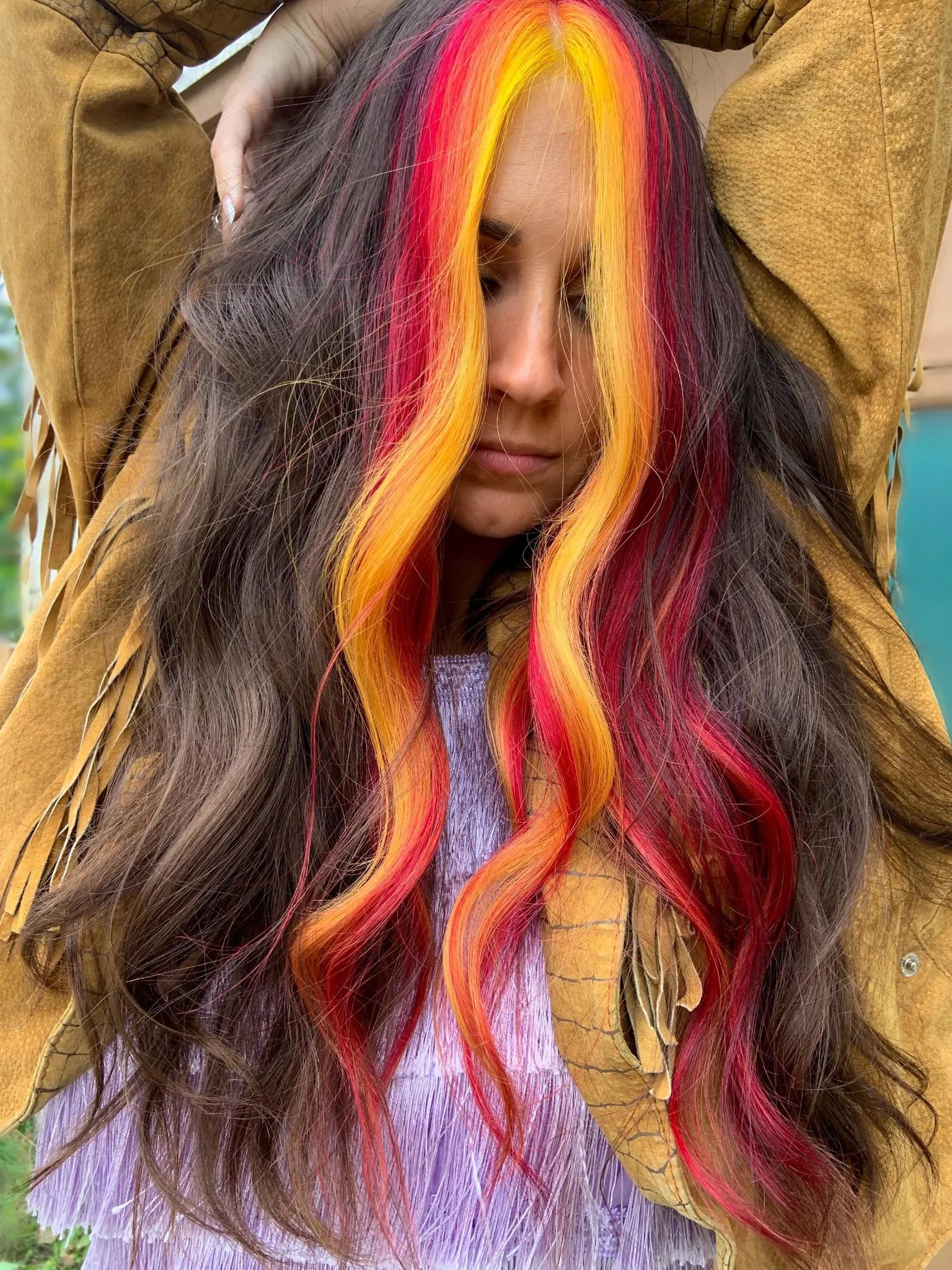A woman with long, wavy hair that is mostly brown with vibrant streaks of red, orange, and yellow. She is wearing a mustard-colored fringed jacket and a lavender top. Her face is partially hidden by her hair and she has her arms raised behind her head.