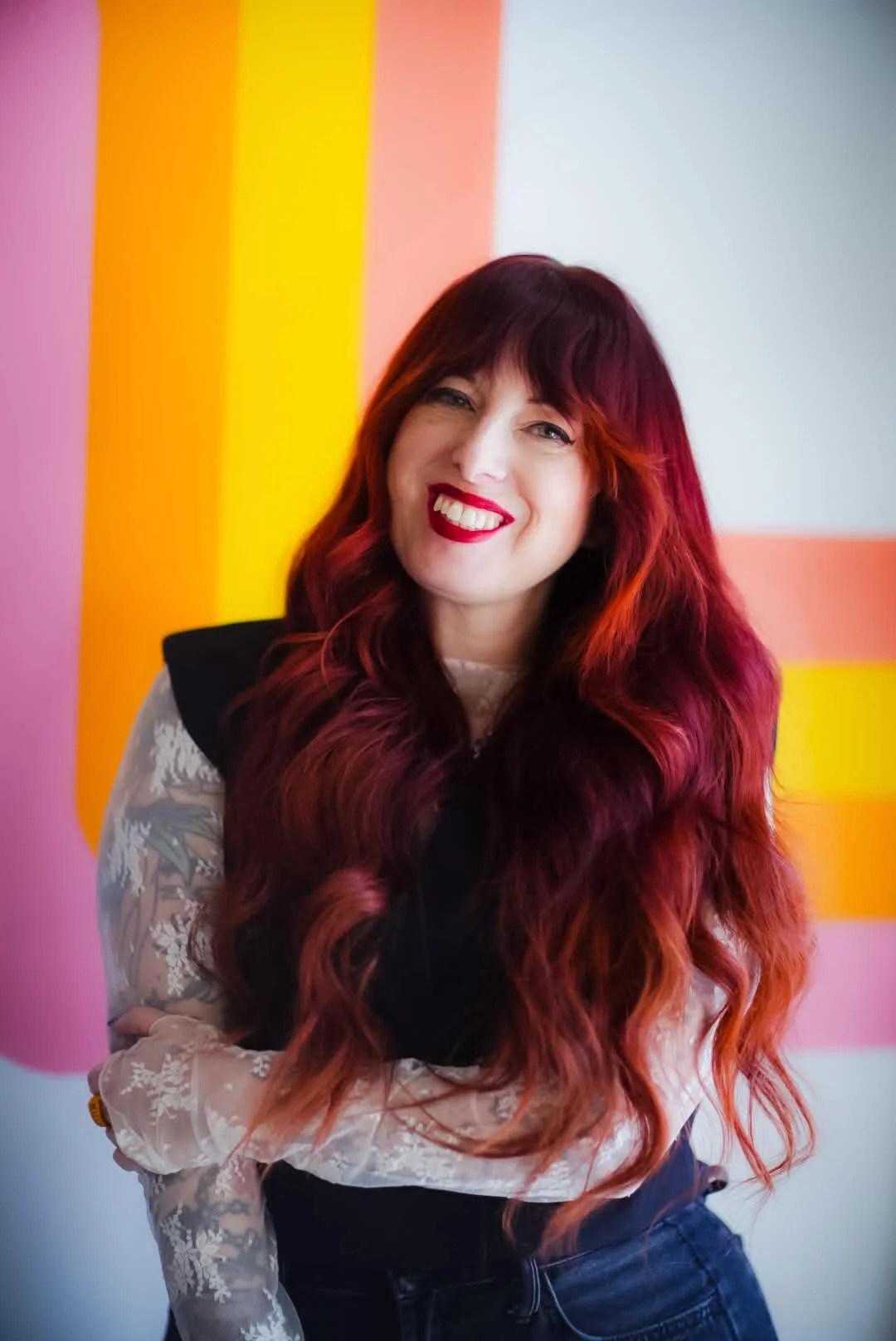 A woman with long red hair and red lipstick smiling in front of a colorful abstract background.