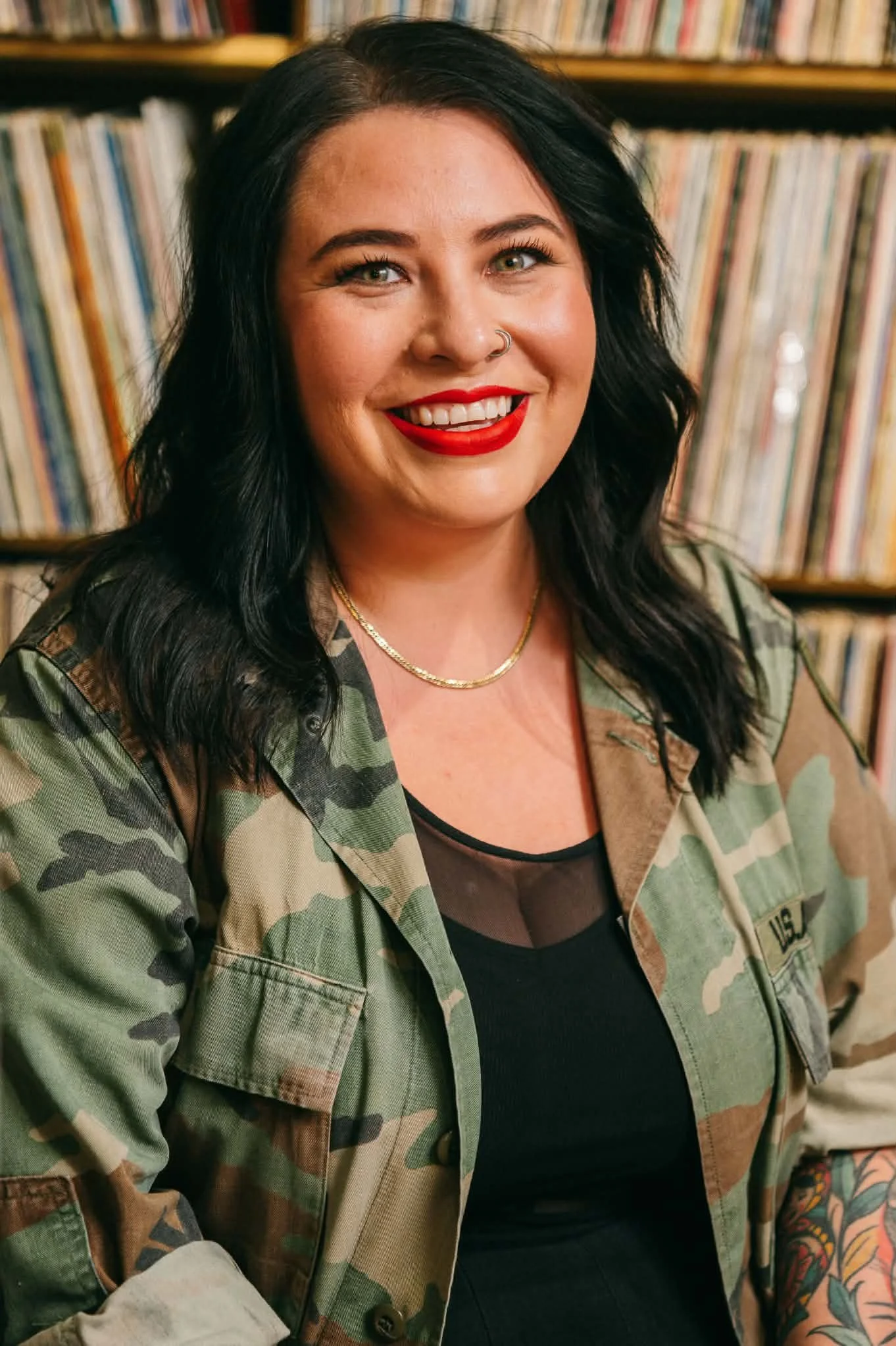 Portrait of a woman with black wavy hair, wearing bright red lipstick, a gold necklace, and a camouflage jacket, in front of a bookshelf filled with records.