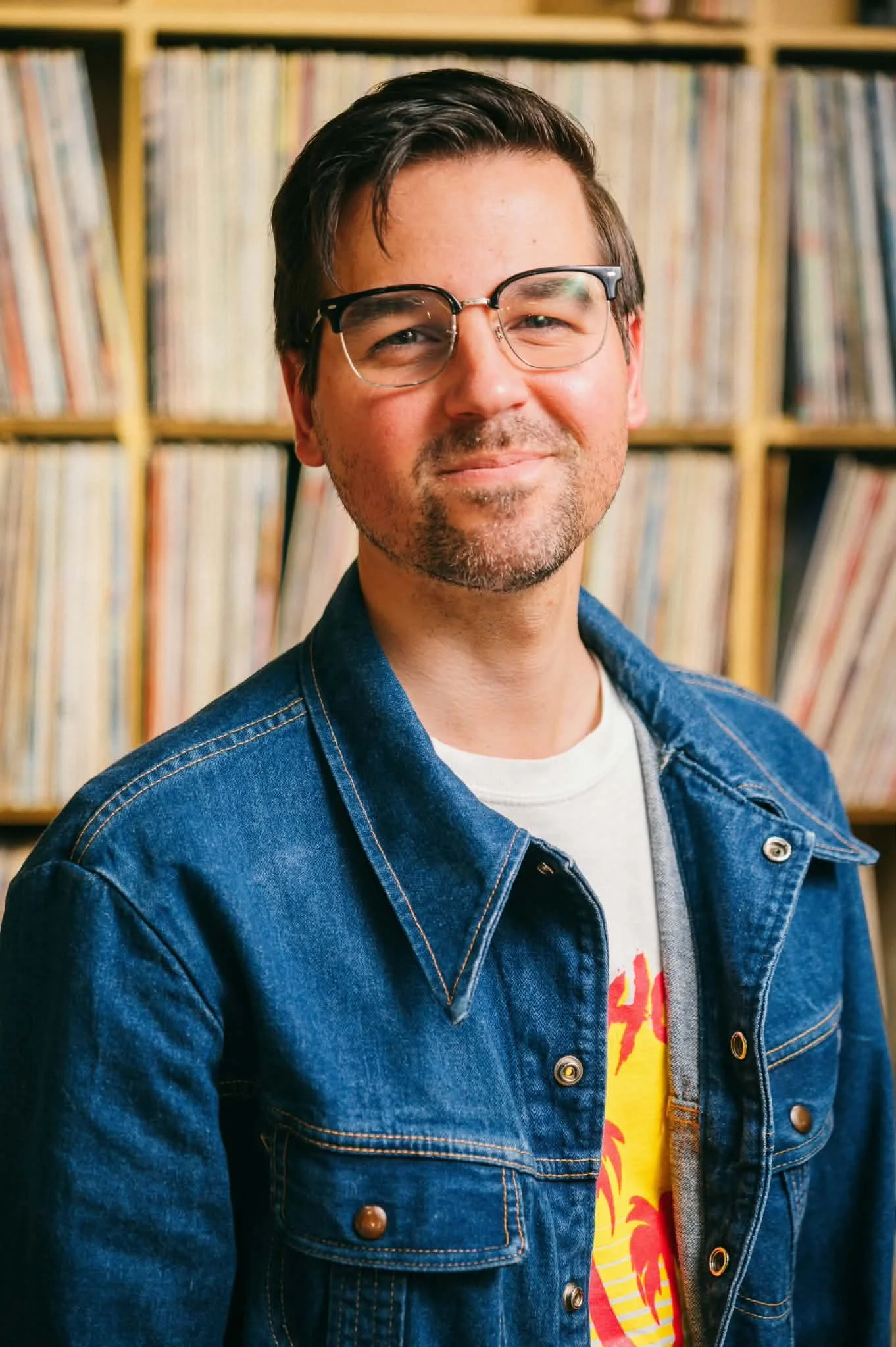 A young man with dark hair, glasses, and a slight beard smiles in front of a bookshelf filled with vinyl records. He is wearing a denim jacket over a colorful graphic t-shirt.