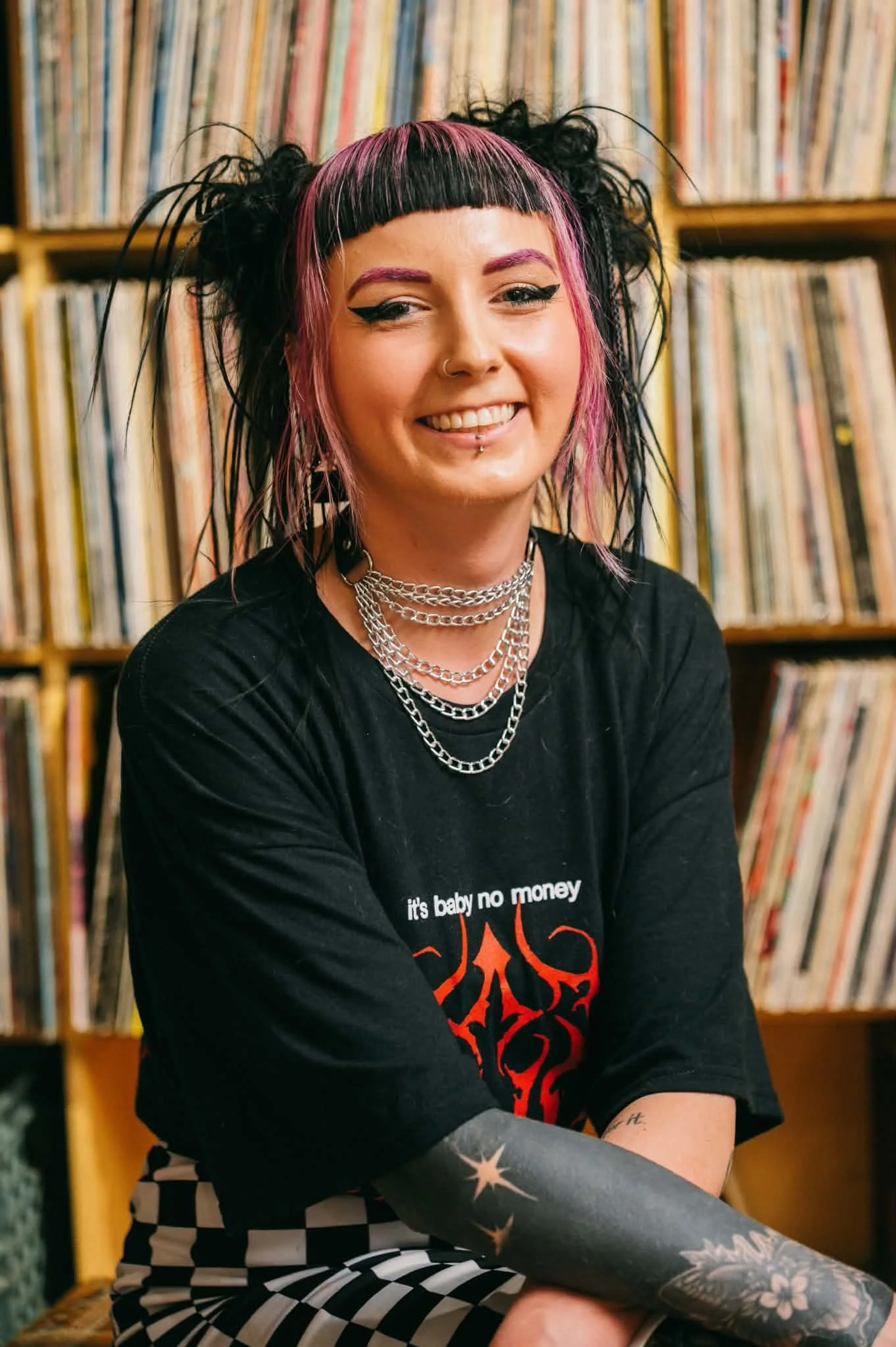 A woman with black and pink hairstyle, wearing multiple silver chains and a black shirt with red graphic, sitting in front of shelves filled with vinyl records.