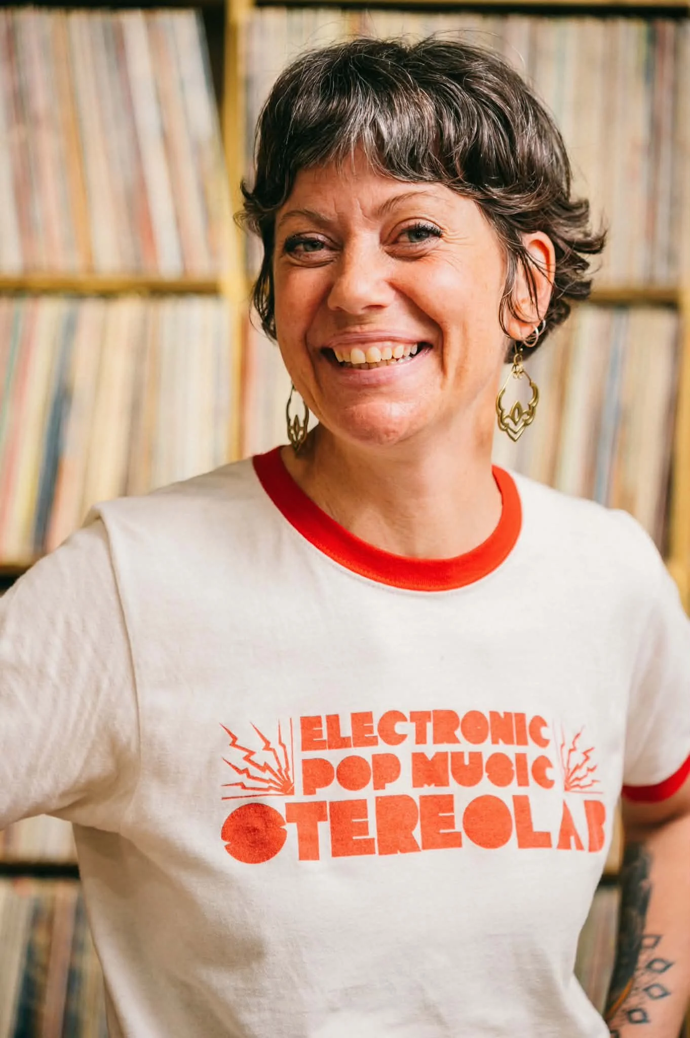 Close-up of a woman with short, dark hair, wearing a white T-shirt with red text, and large yellow earrings. She is smiling while standing in front of a bookshelf filled with vinyl records.