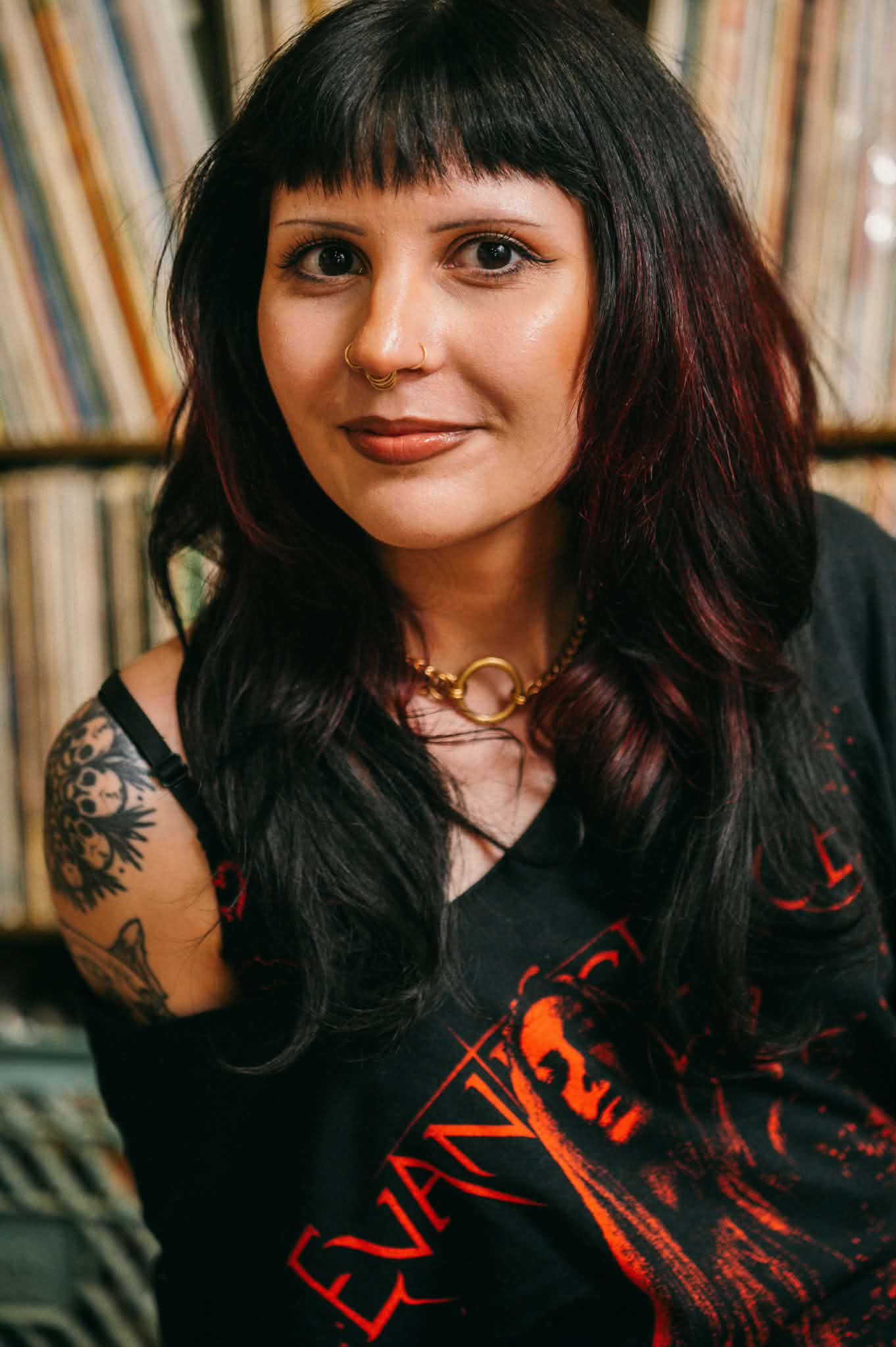 A woman with long black hair with red highlights, tattoos on her left arm, and wearing a black t-shirt, standing in front of a bookshelf filled with vinyl records.