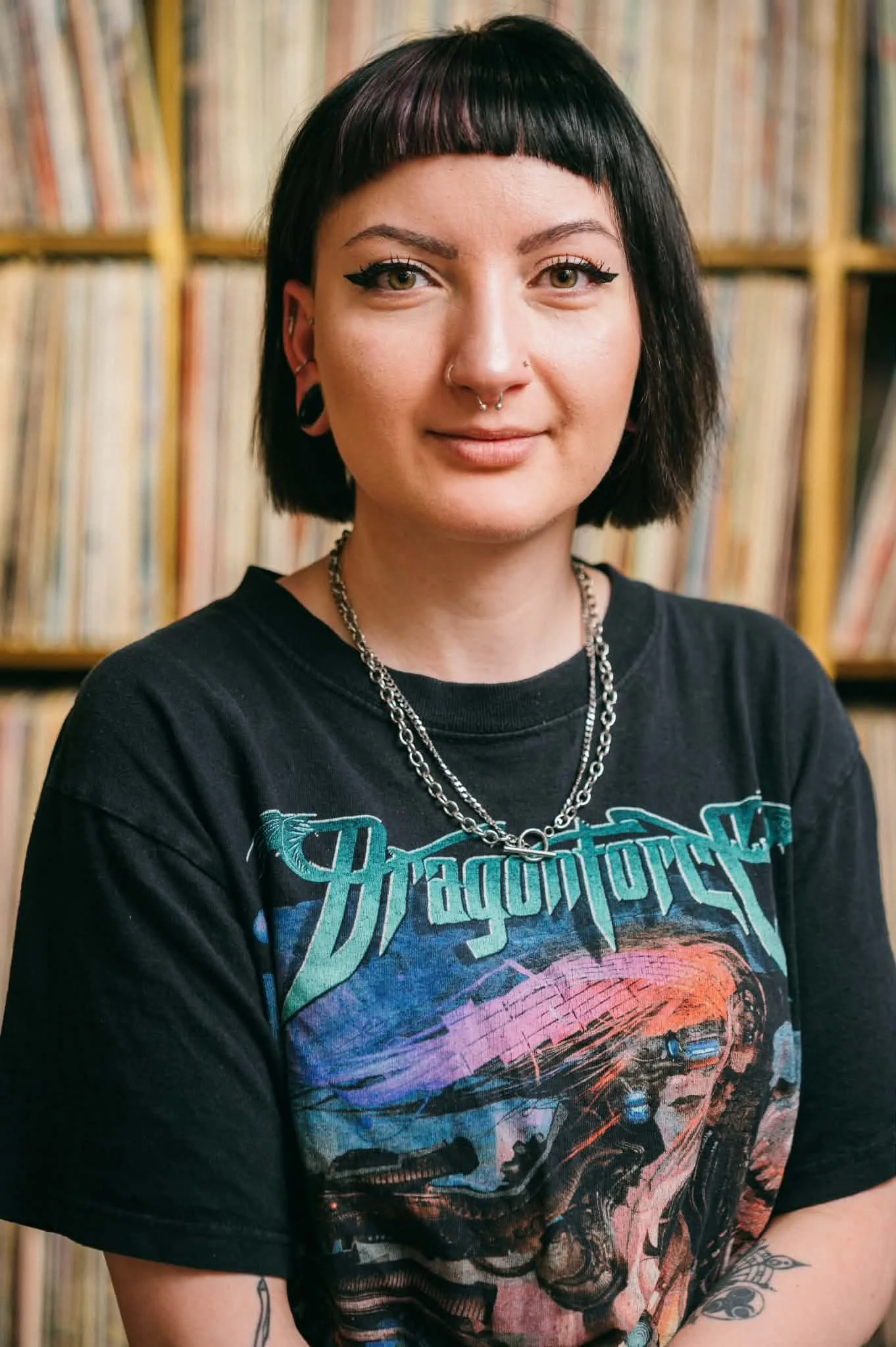 Portrait of a young woman with black hair, tattoos, and piercings, standing in front of a bookshelf filled with vinyl records.