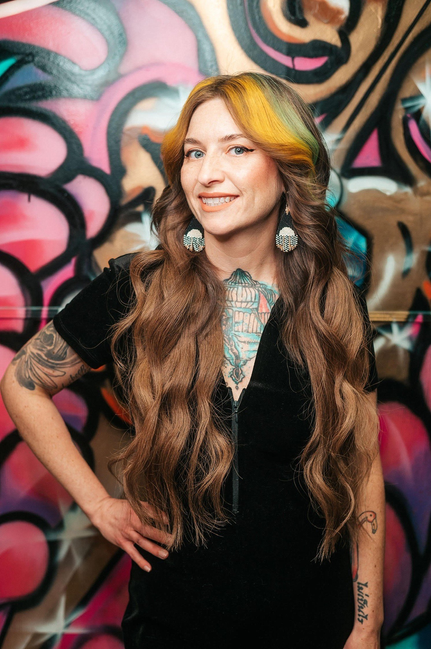 A woman with long wavy hair with blonde and green streaks, wearing large fan-shaped earrings, standing in front of a colorful graffiti wall with pink, black, and brown abstract designs.