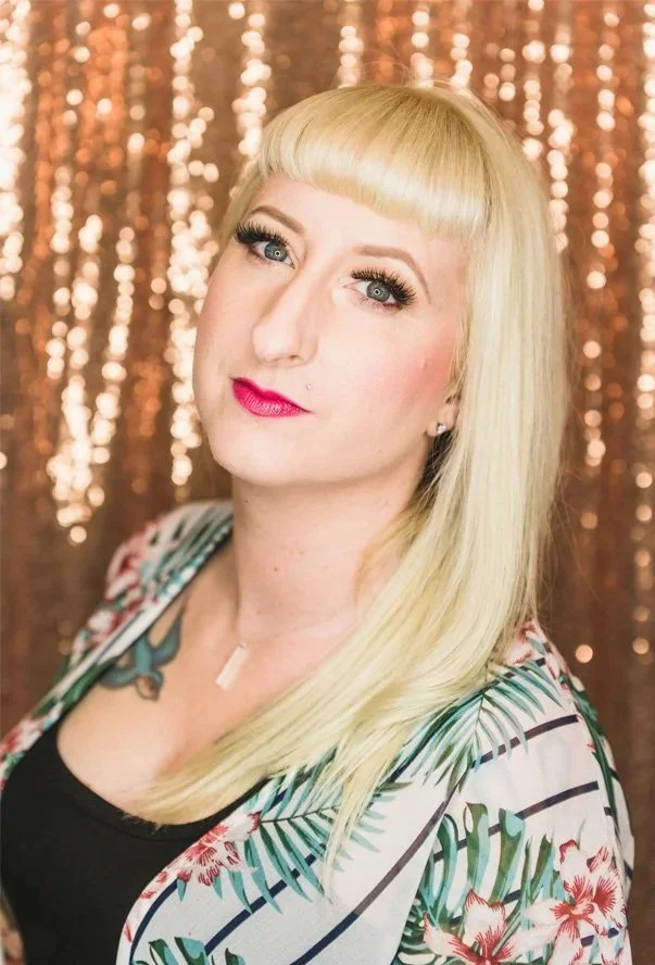 A woman with blonde hair, wearing makeup including red lipstick and eye makeup, standing in front of a shimmering gold backdrop, wearing a black top and a floral patterned jacket with green leaves and pink flowers.