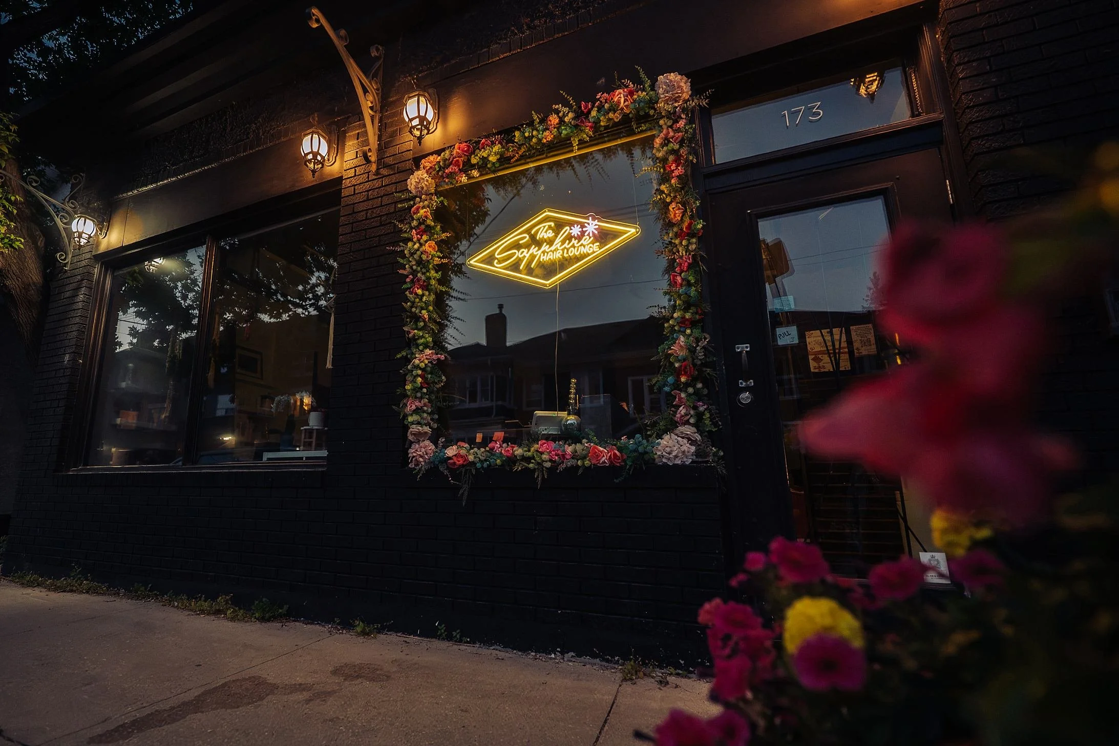 Night view of a hair salon storefront with illuminated neon sign "The Sapphire Hair Lounge" surrounded by a floral wreath, black brick exterior, and decorative outdoor lamps.