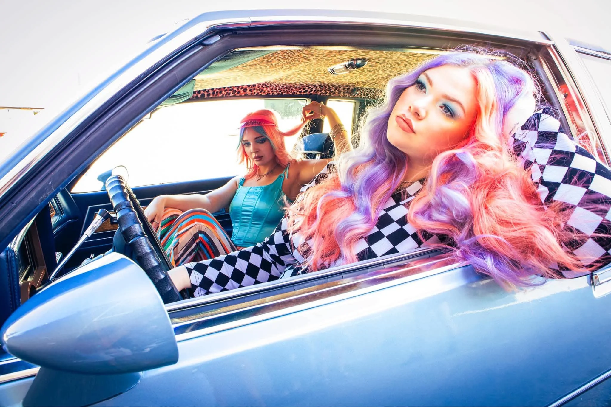 Two women with colorful hair and vibrant makeup sitting inside a vintage blue car, with a leopard print and checkered pattern on the ceiling, showcasing a fashion-forward, retro style.
