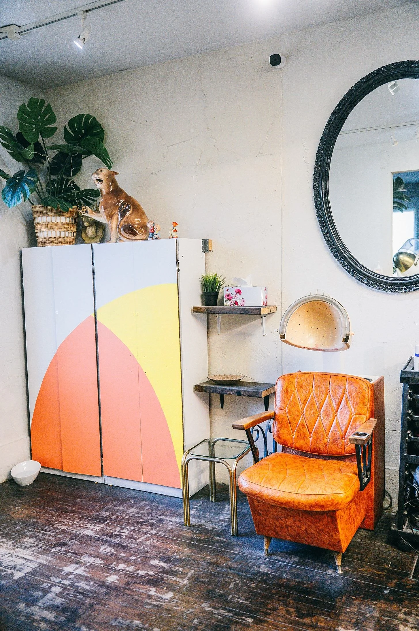 A cozy room corner with a vintage orange armchair, a small metal and wood side table, a wall mirror with a black ornate frame, a decorative ceramic dog statue, greenery in a planter, and a colorful geometric room divider.