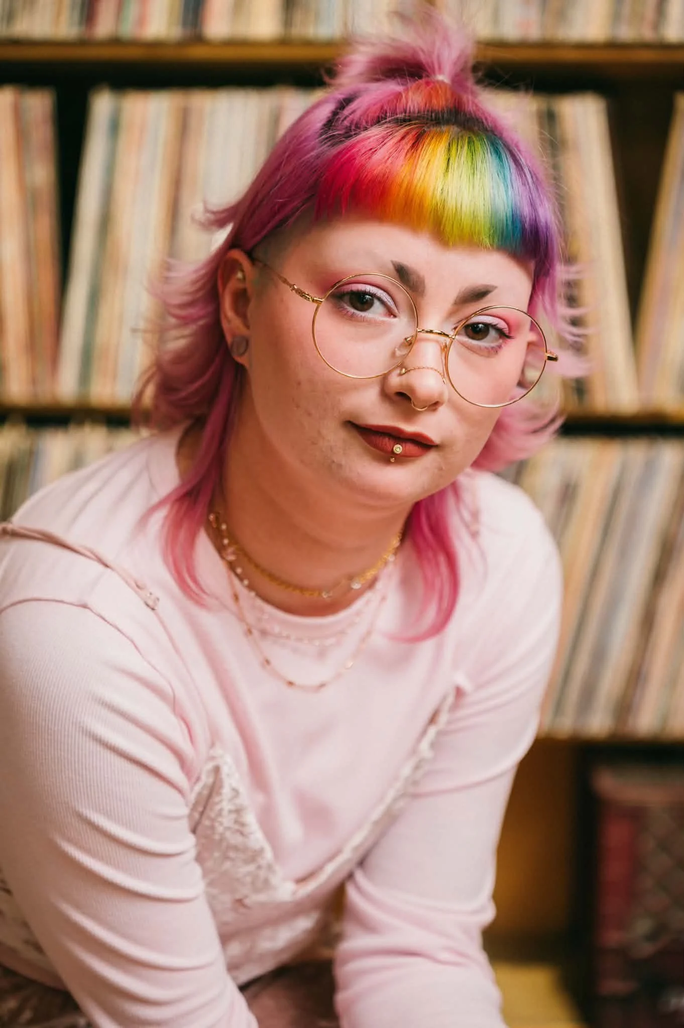 A person with pink hair, colorful rainbow bangs, and wearing round glasses and gold jewelry poses in front of a bookshelf filled with vinyl records.