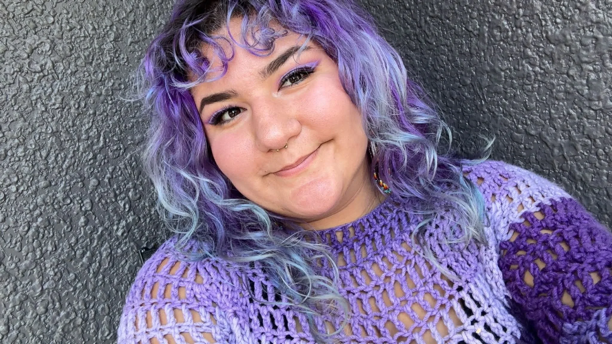 Smiling woman with multicolored blue and purple hair, wearing a black top and gold earrings, standing in front of a graffiti mural with pink, black, and blue designs.