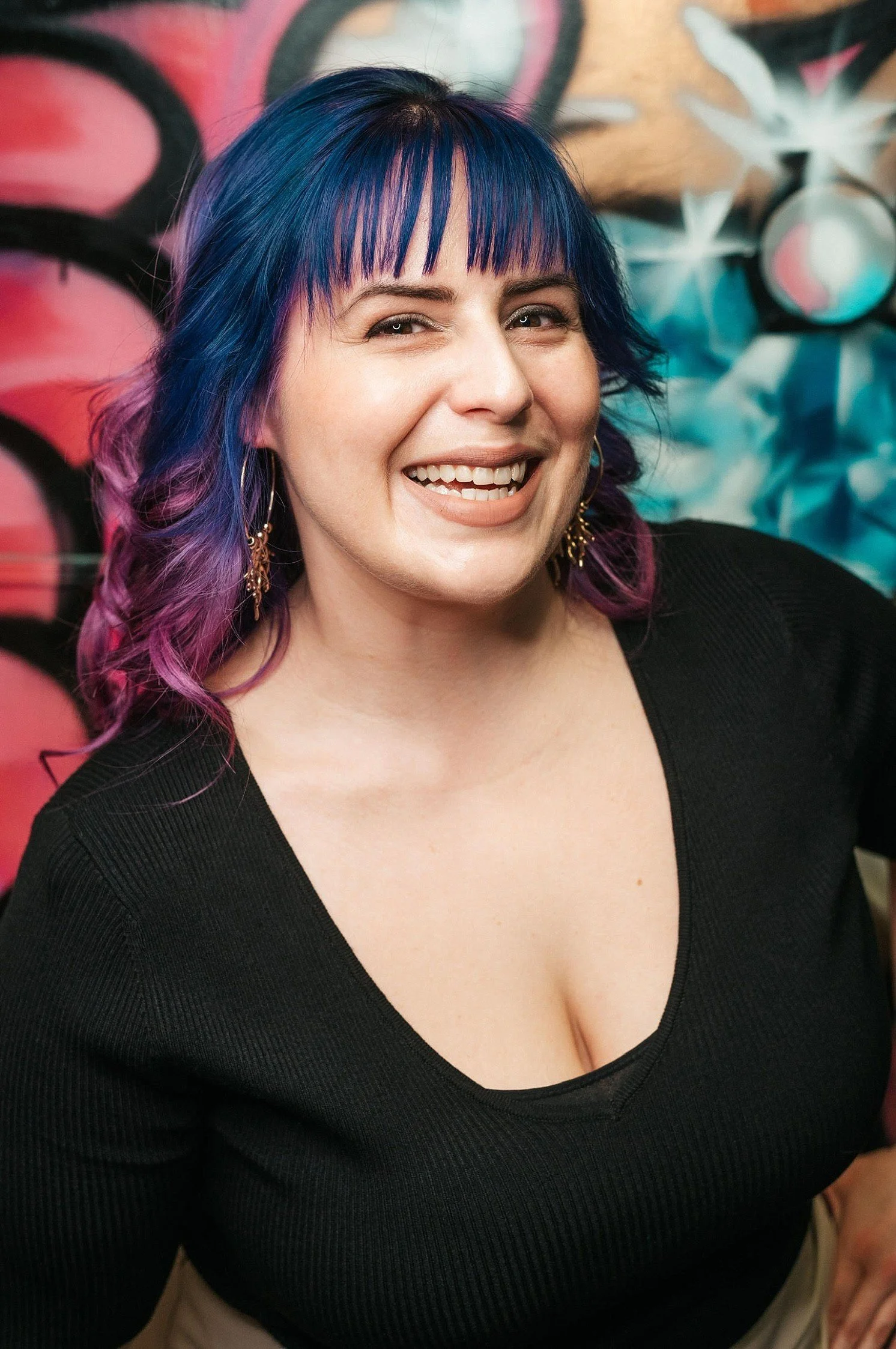 Smiling woman with multicolored blue and purple hair, wearing a black top and gold earrings, standing in front of a graffiti mural with pink, black, and blue designs.