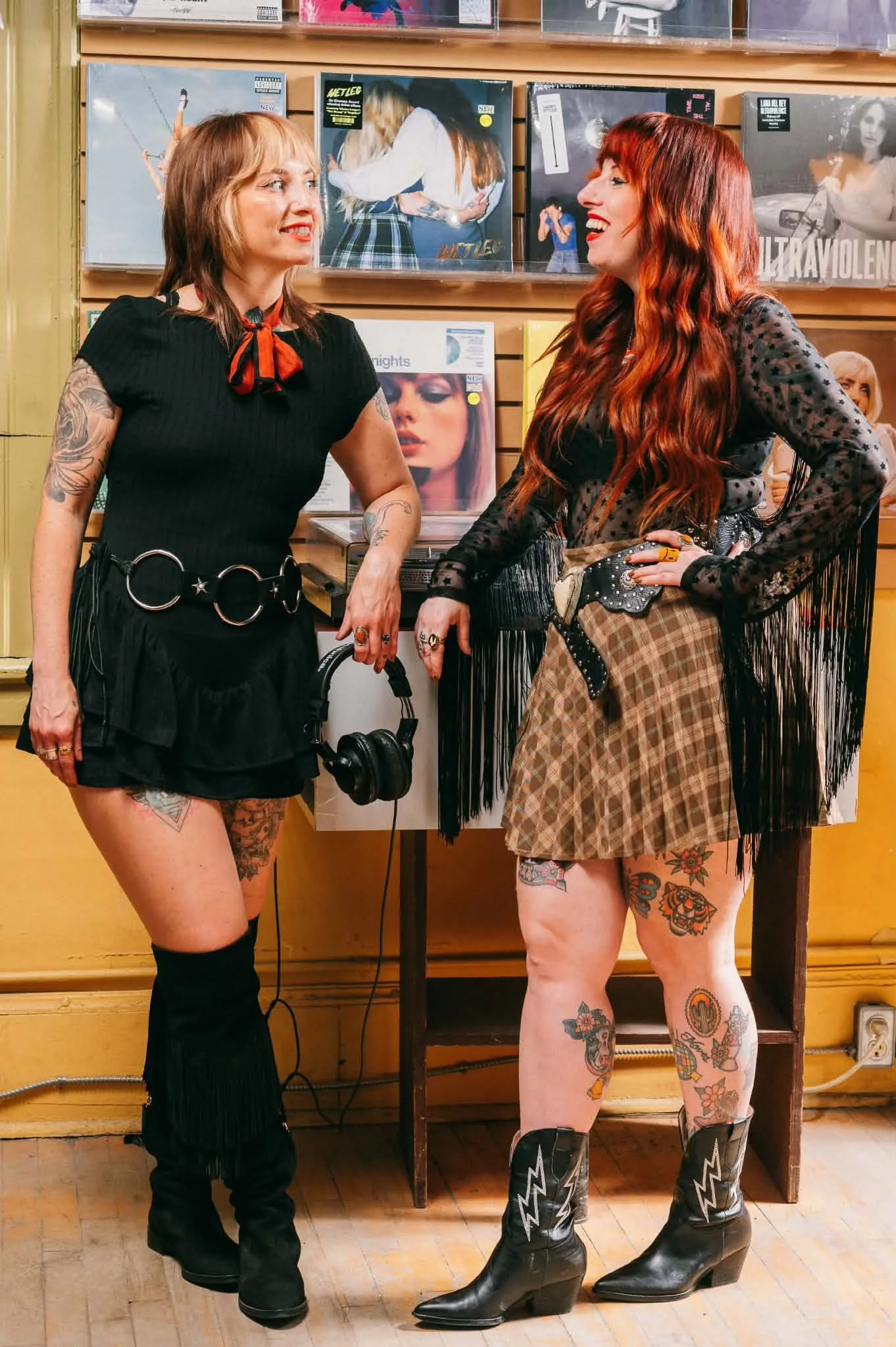 Two women with tattoos and colorful hair stand in a record store, smiling and facing each other. One woman is wearing a black dress with a red scarf, knee-high black boots, and is holding headphones. The other woman is dressed in a black lace top, plaid skirt, ripped shorts, cowboy boots with lightningbolt designs, and has tattoos on her legs. They are standing in front of a wall with vinyl records.
