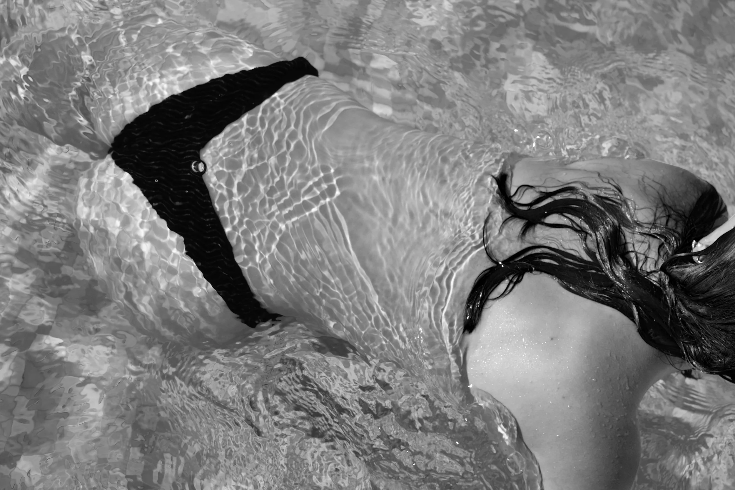 A person with long dark hair submerged in water, wearing a black long-sleeve shirt, with part of their face and shoulder visible, and ripples in the water surrounding them.