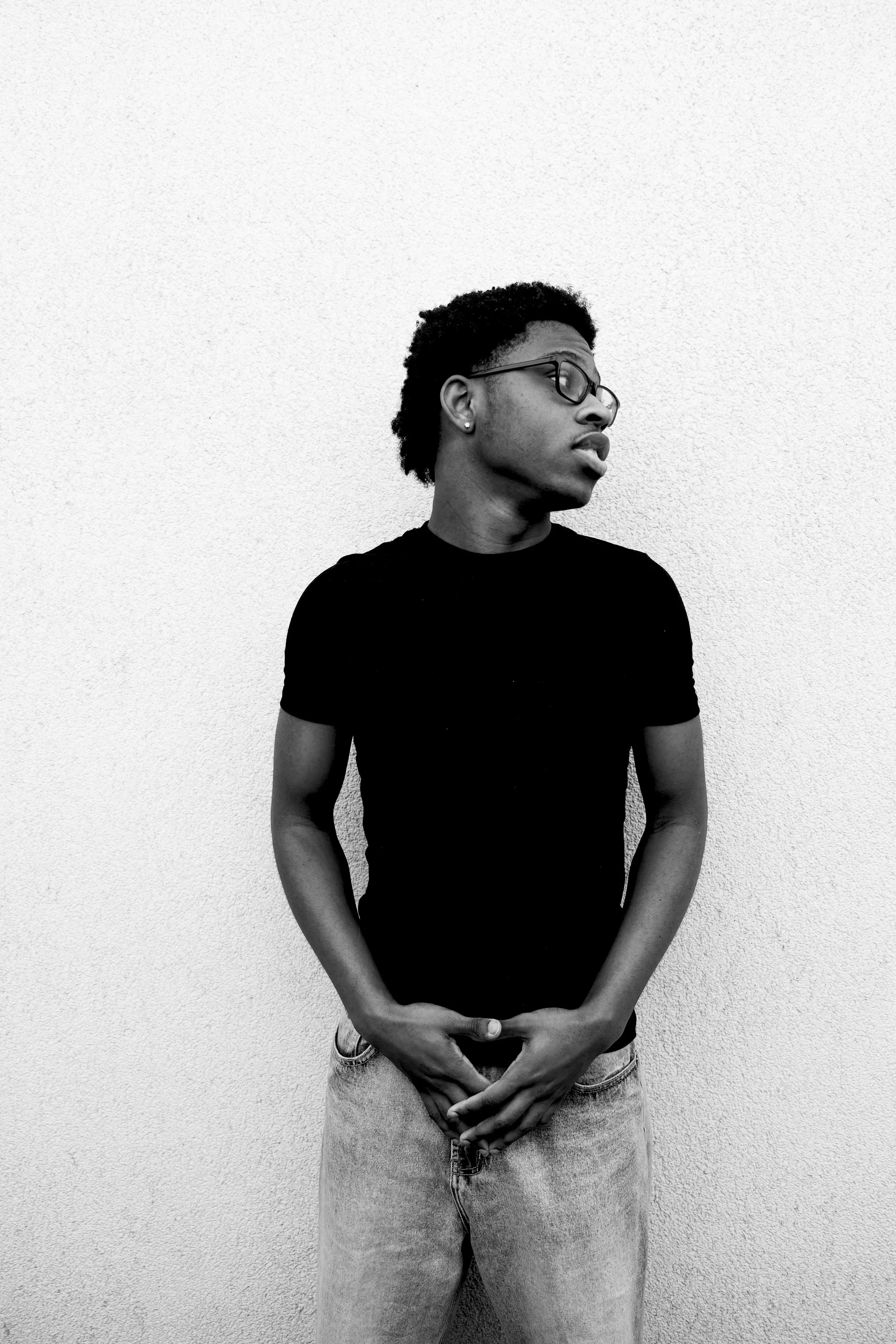Black and white photo of a young man wearing glasses, a black t-shirt, and jeans, standing against a plain textured wall, with his head turned to the side and his hands clasped in front.