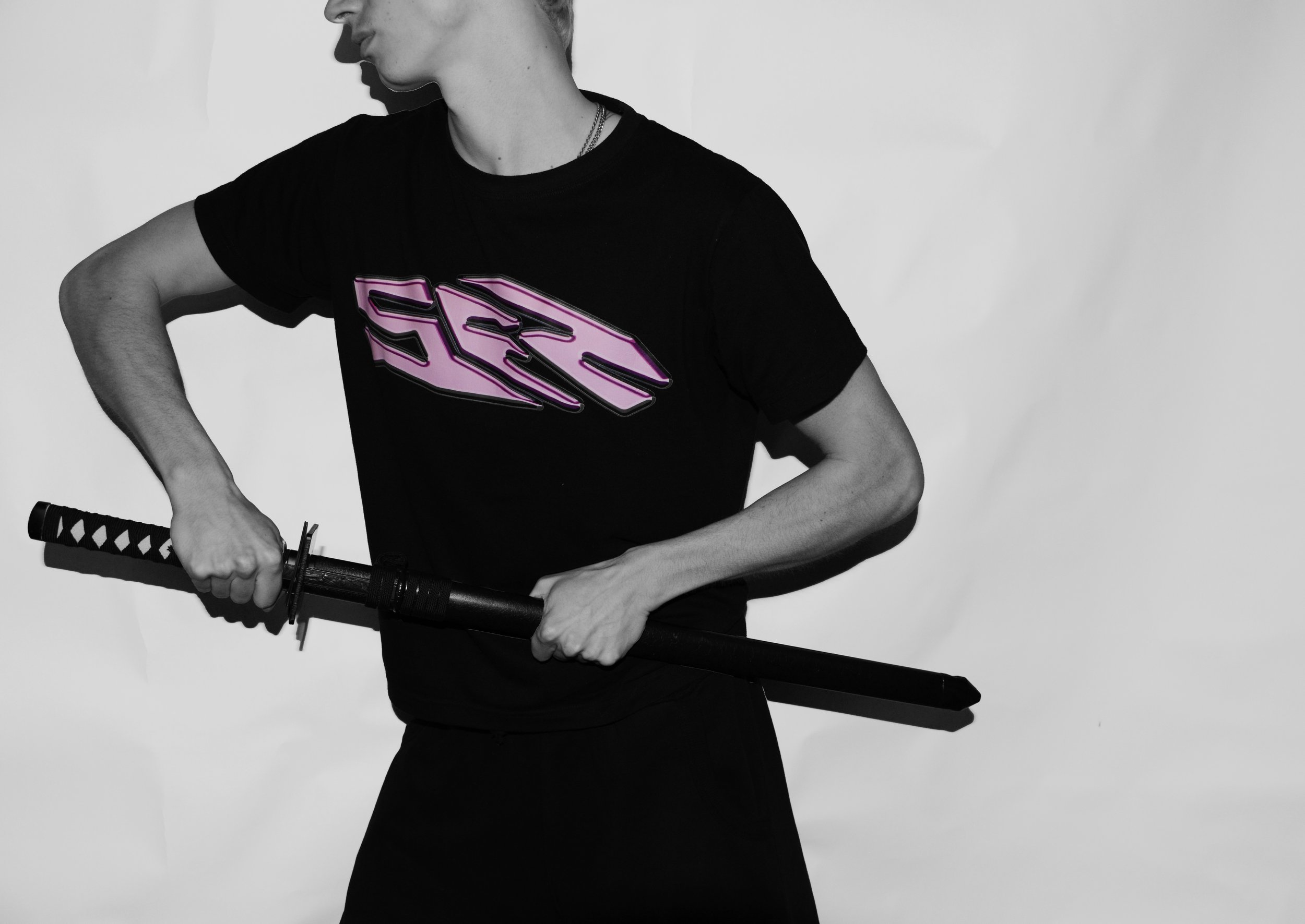 Person holding a katana sword with a black and white background. The person is wearing a black t-shirt with a pink geometric design, and has a silver chain around their neck.