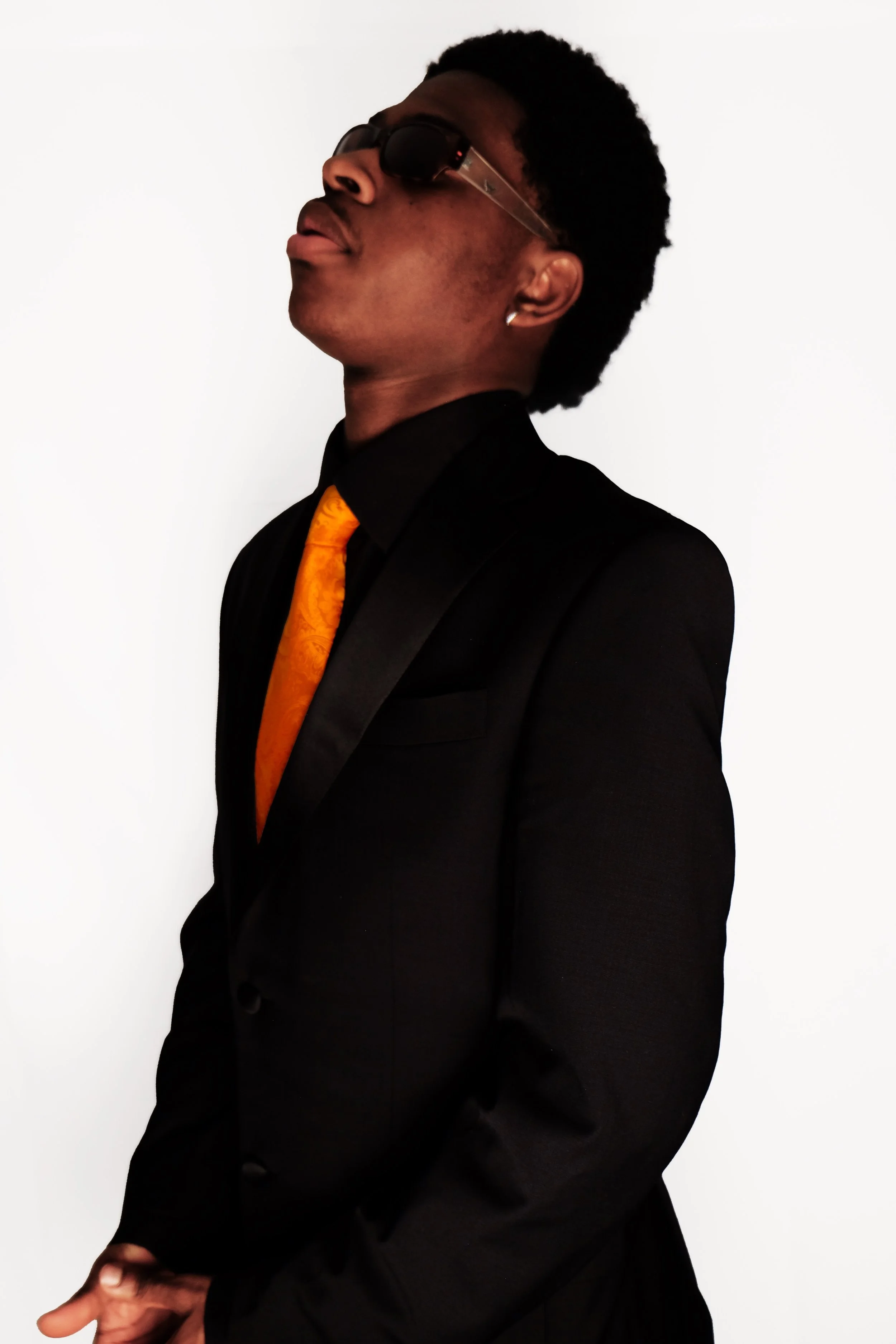 A young man in a black suit, orange tie, and sunglasses, standing in profile against a plain white background.