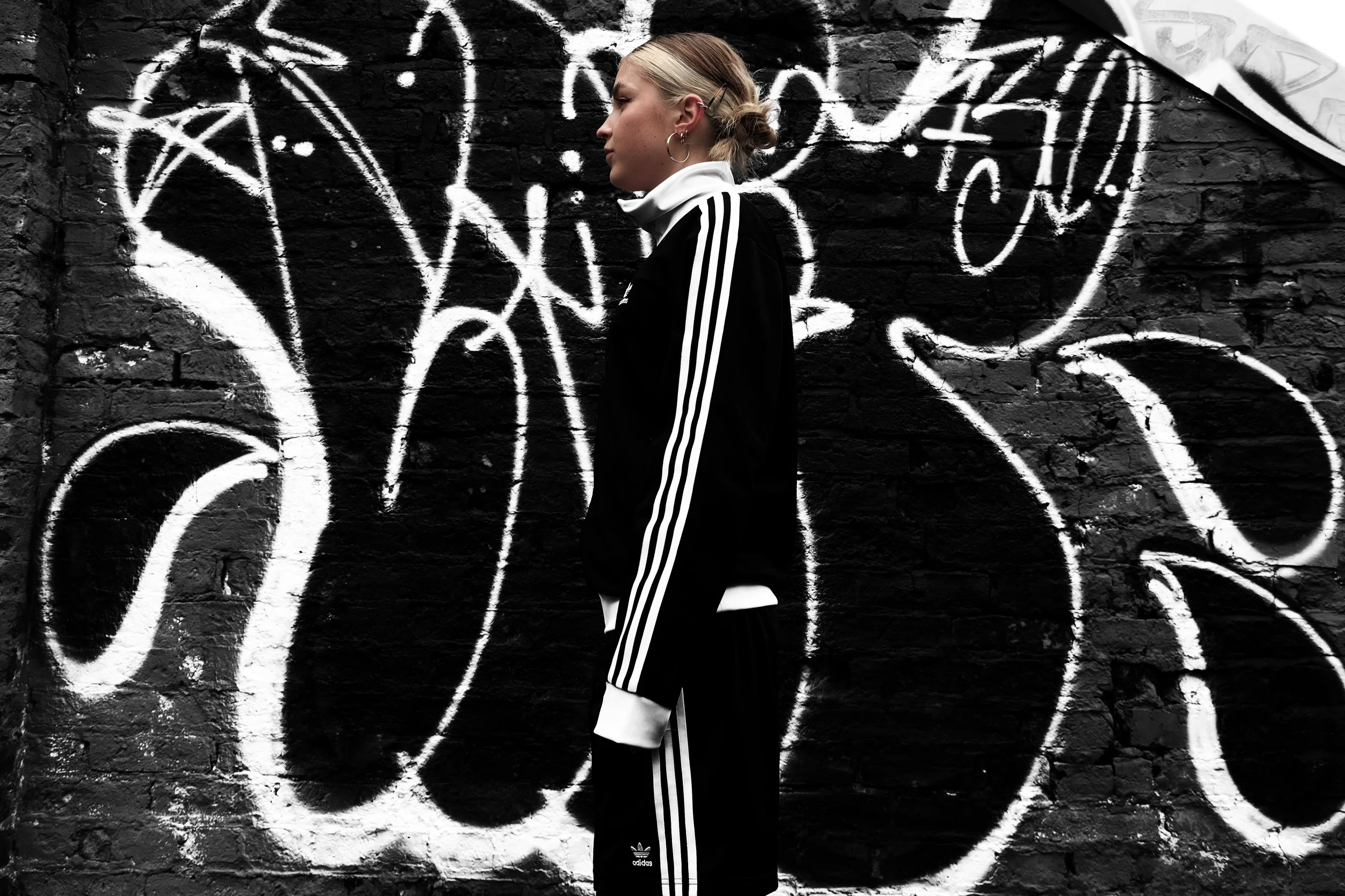 A woman with light blonde hair tied back with a clip, dressed in a black Adidas tracksuit with white stripes, standing sideways against a black brick wall with white graffiti art.