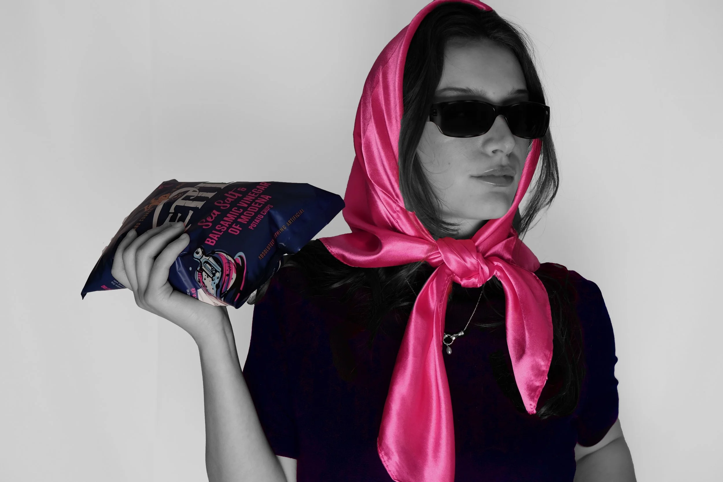 A woman with long dark hair wearing sunglasses and a vibrant pink satin scarf tied around her head, holding a bag of salt over her shoulder, captured in black and white with the exception of the pink scarf and salt bag which are in color.