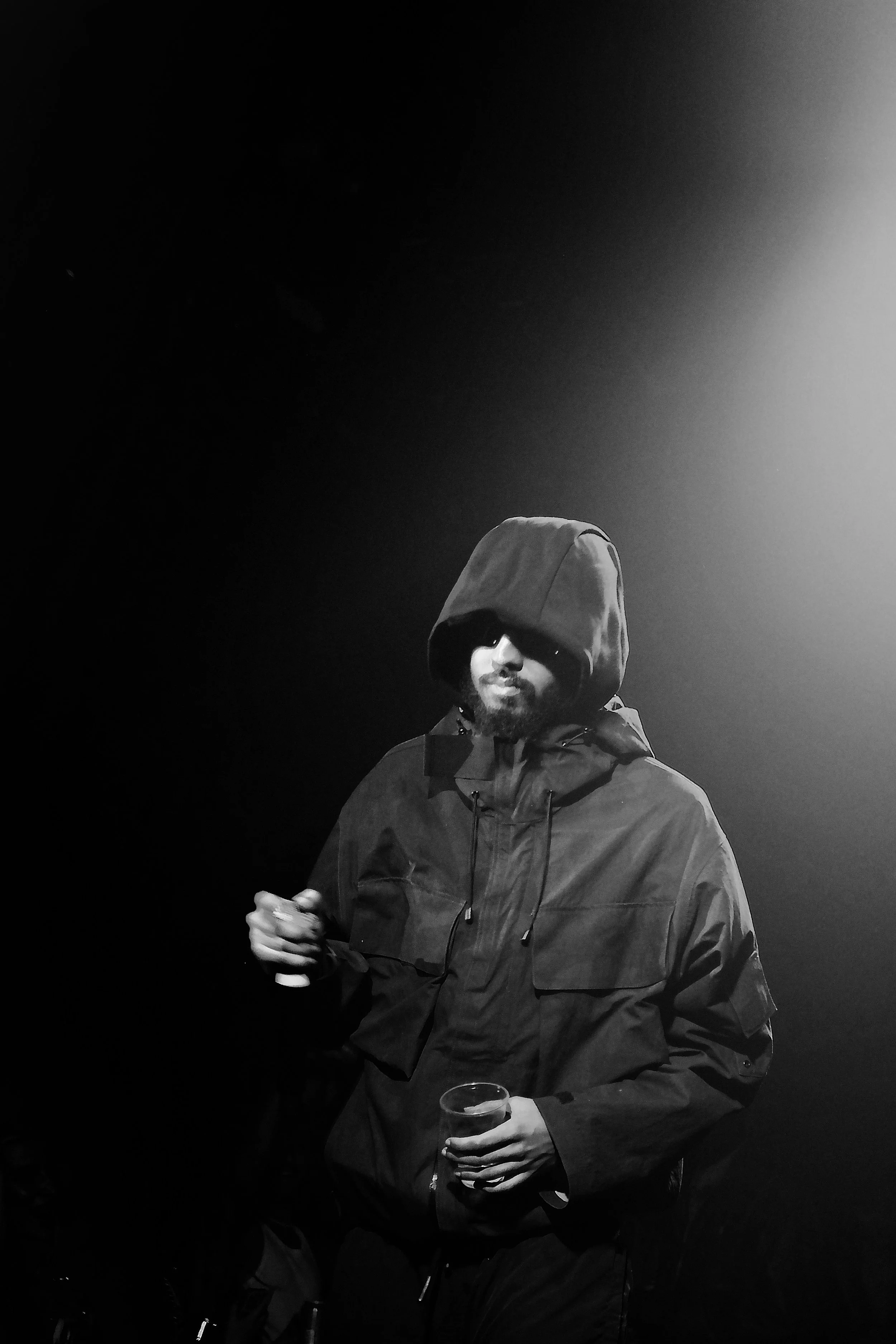 A man wearing a hoodie and sunglasses, holding a drink in one hand and a phone in the other, in a dark environment with dramatic lighting.