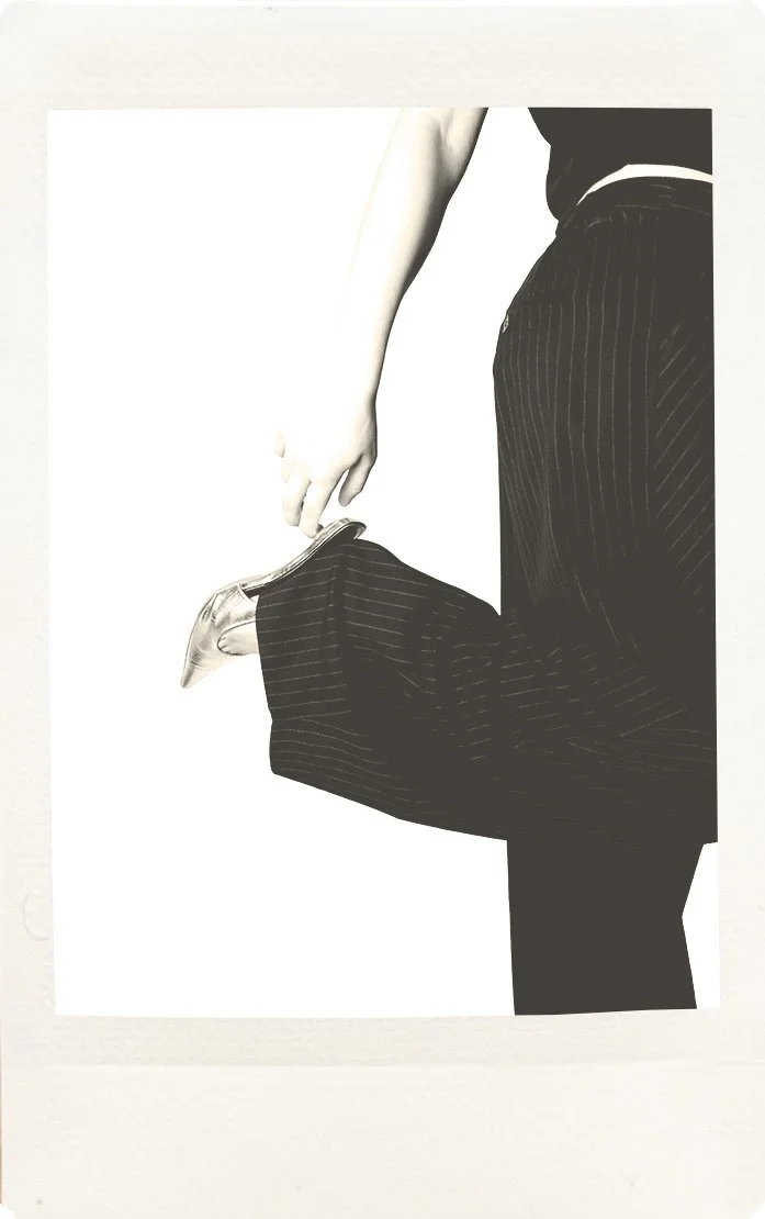 A person in a business suit pulling up their pant leg to reveal a shiny high-heeled shoe.