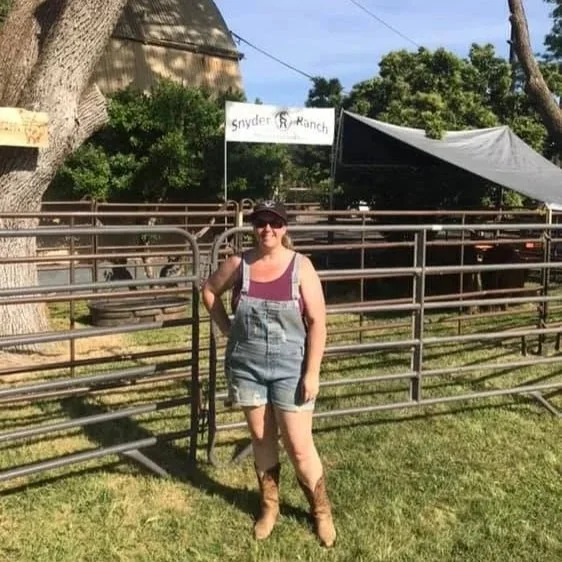 A woman stands outdoors on grass, wearing sunglasses, a hat, tank top, denim overalls, and cowboy boots, with a cattle pen and a sign that reads 'Snyder Ranch' in the background.