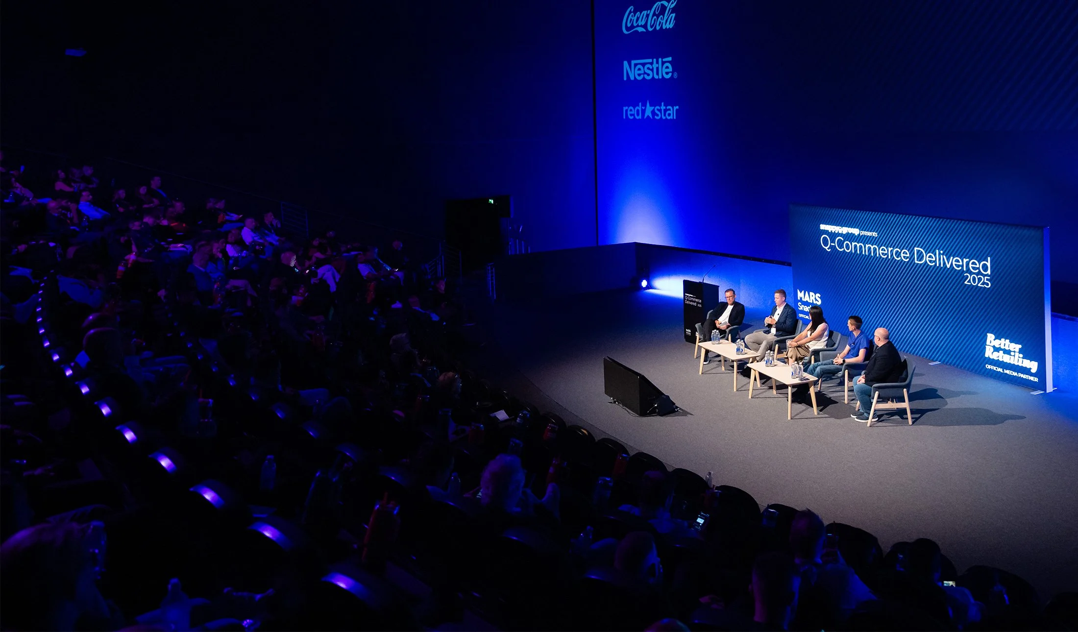Conference stage with panel of speakers, large screen displaying event title 'Q-Commerce Delivered 2025' and logos of Coca-Cola, Nestlé, Red Star. Audience seated in dark auditorium.