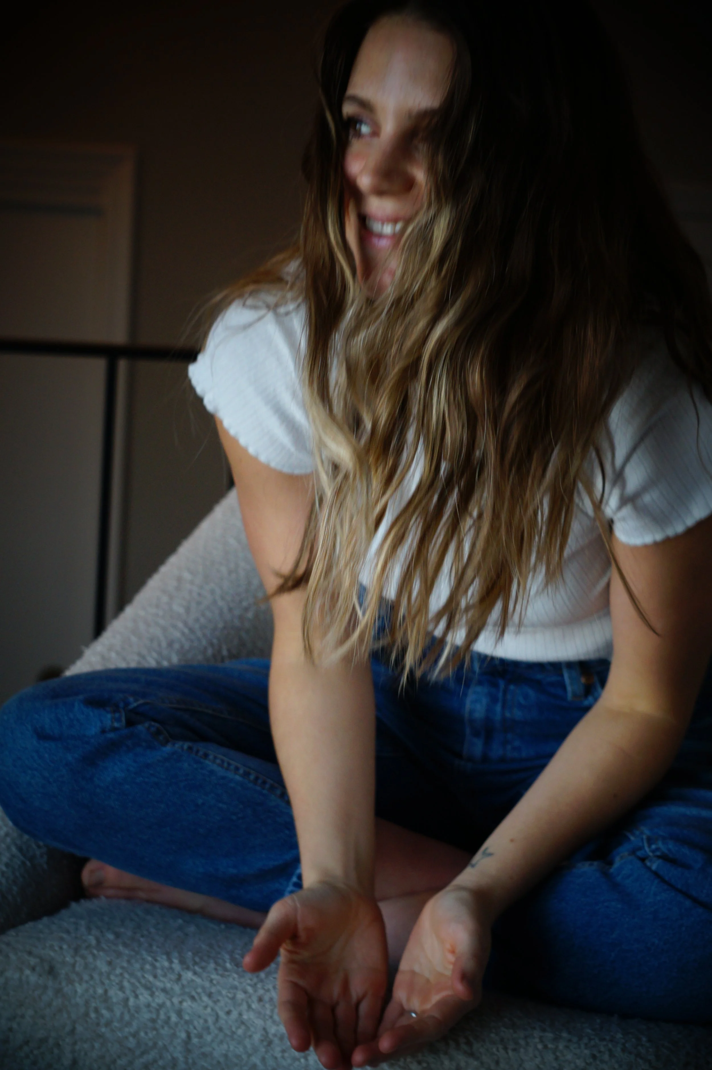 A woman with long wavy hair sitting cross-legged on a carpeted floor, smiling and looking to her left, wearing a white t-shirt and blue jeans.