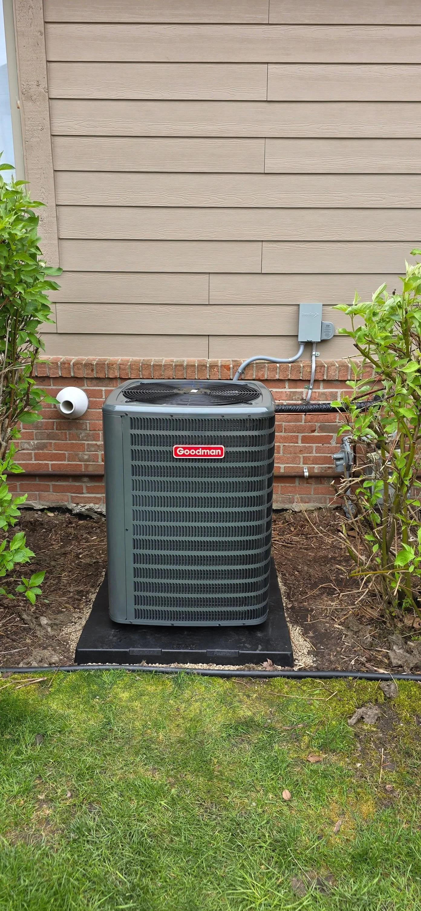 Central air conditioning unit outside a house, surrounded by small bushes and grass, with a brick foundation and beige siding on the house wall.