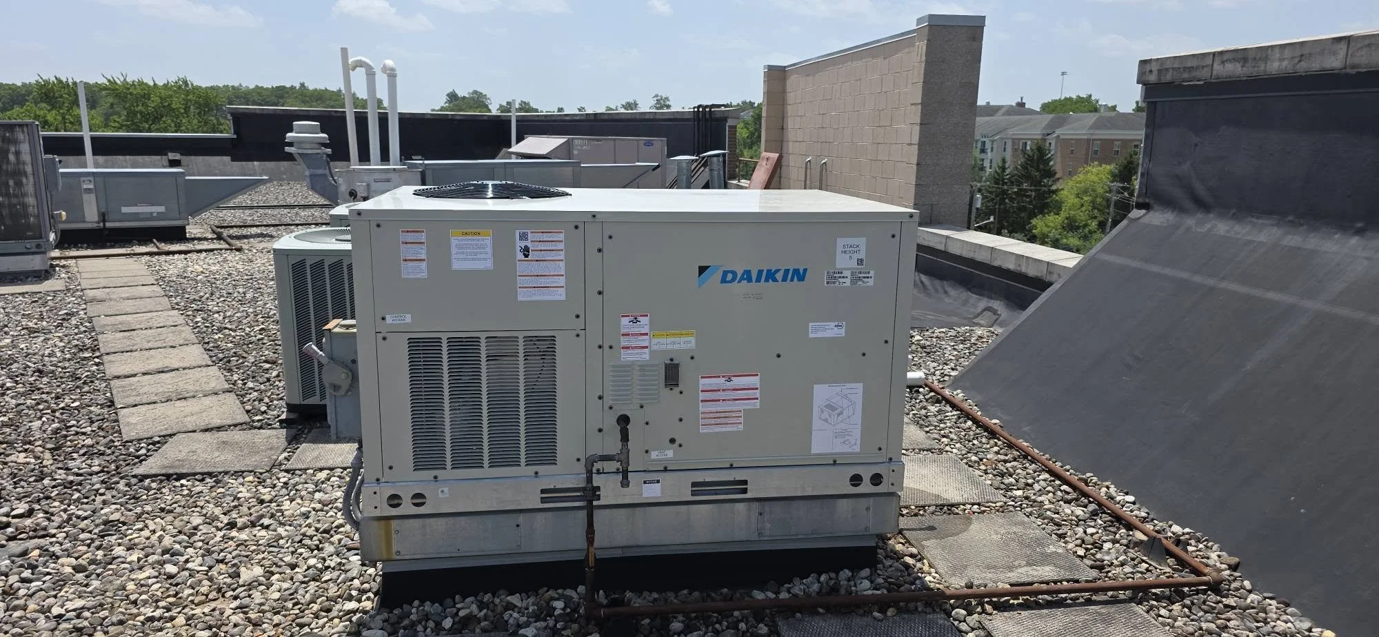 On a building rooftop, a large beige Daikin HVAC unit sits on a gravel surface, surrounded by ventilation equipment and piping, with trees and buildings in the background.