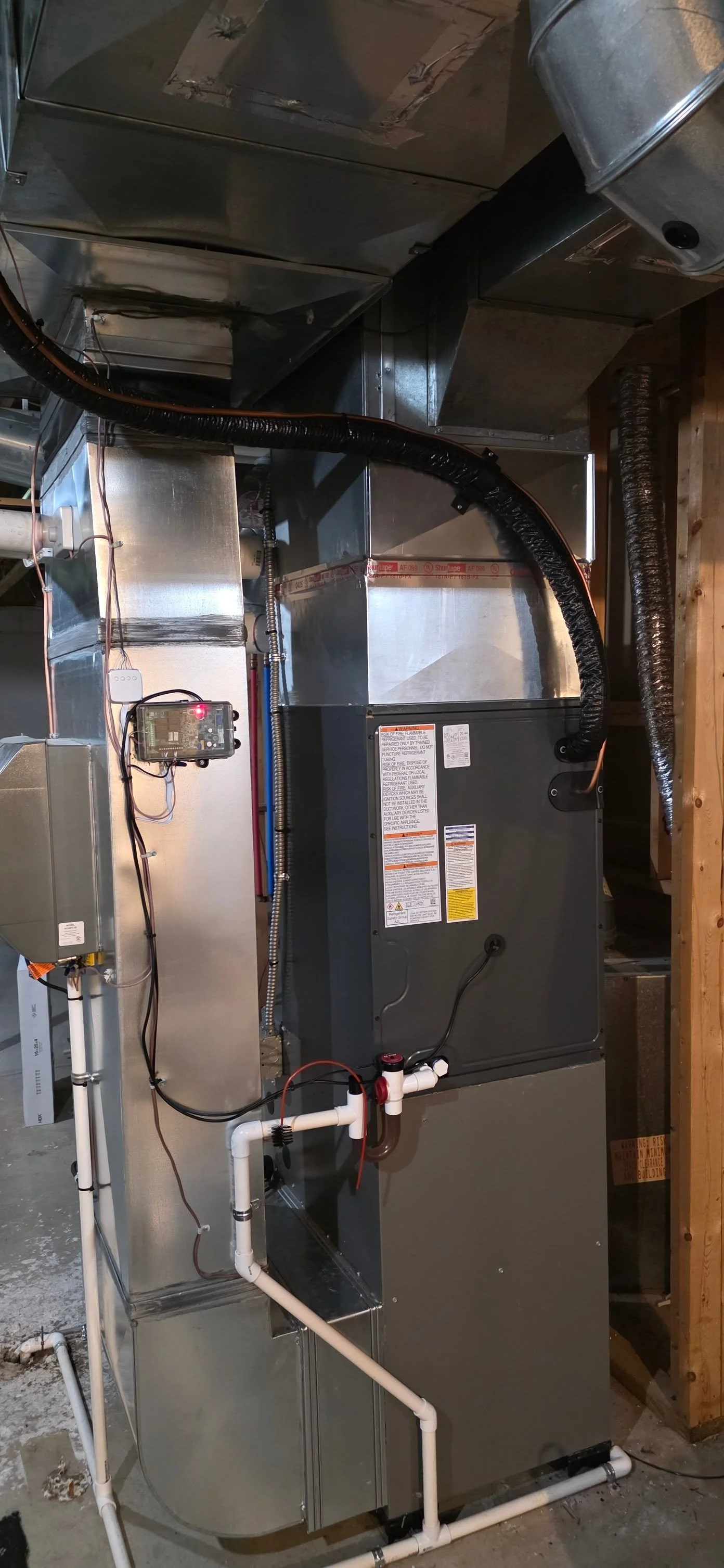 A large HVAC furnace unit with metal vents, pipes, wiring, and a control board installed in a basement or utility room.