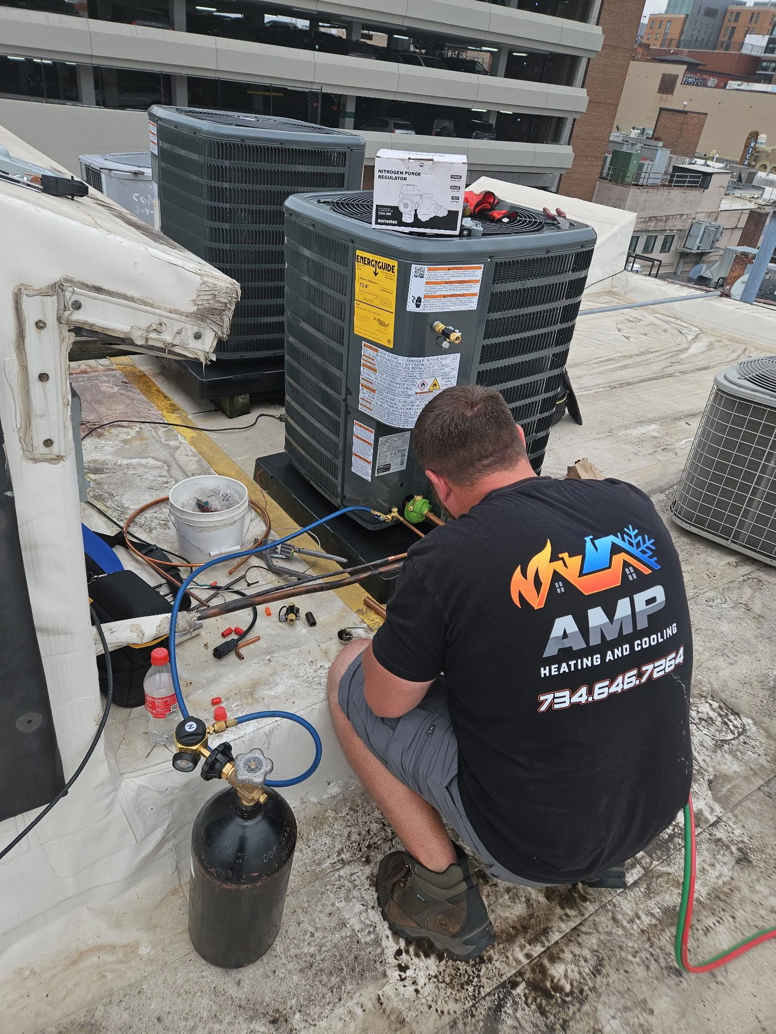 A technician is kneeling on a rooftop, working on an air conditioning unit connected to a nitrogen purge regulator, with various tools and equipment around him.