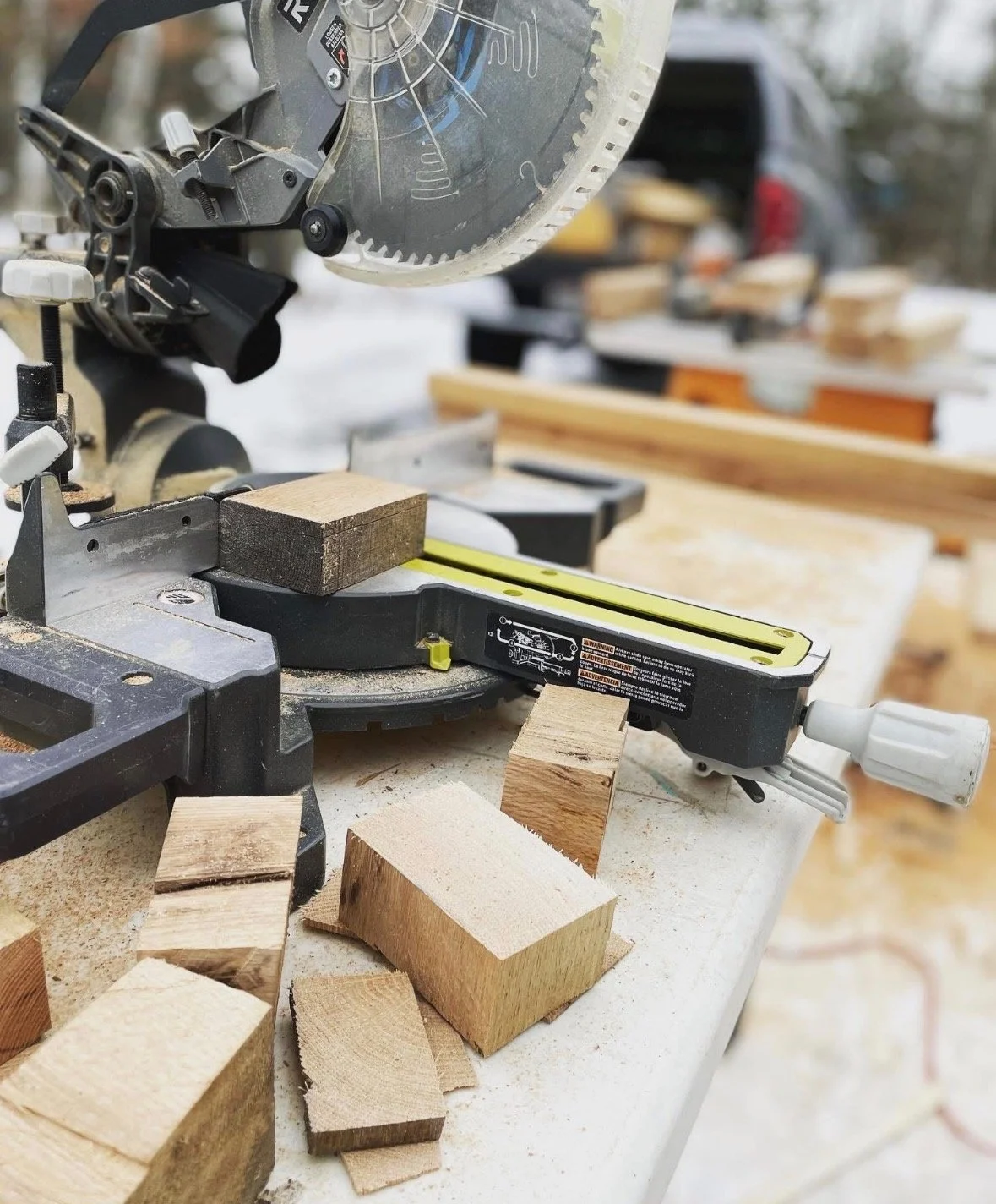 A miter saw cutting a piece of wood outdoors, surrounded by wood scraps and more wood pieces in the background.