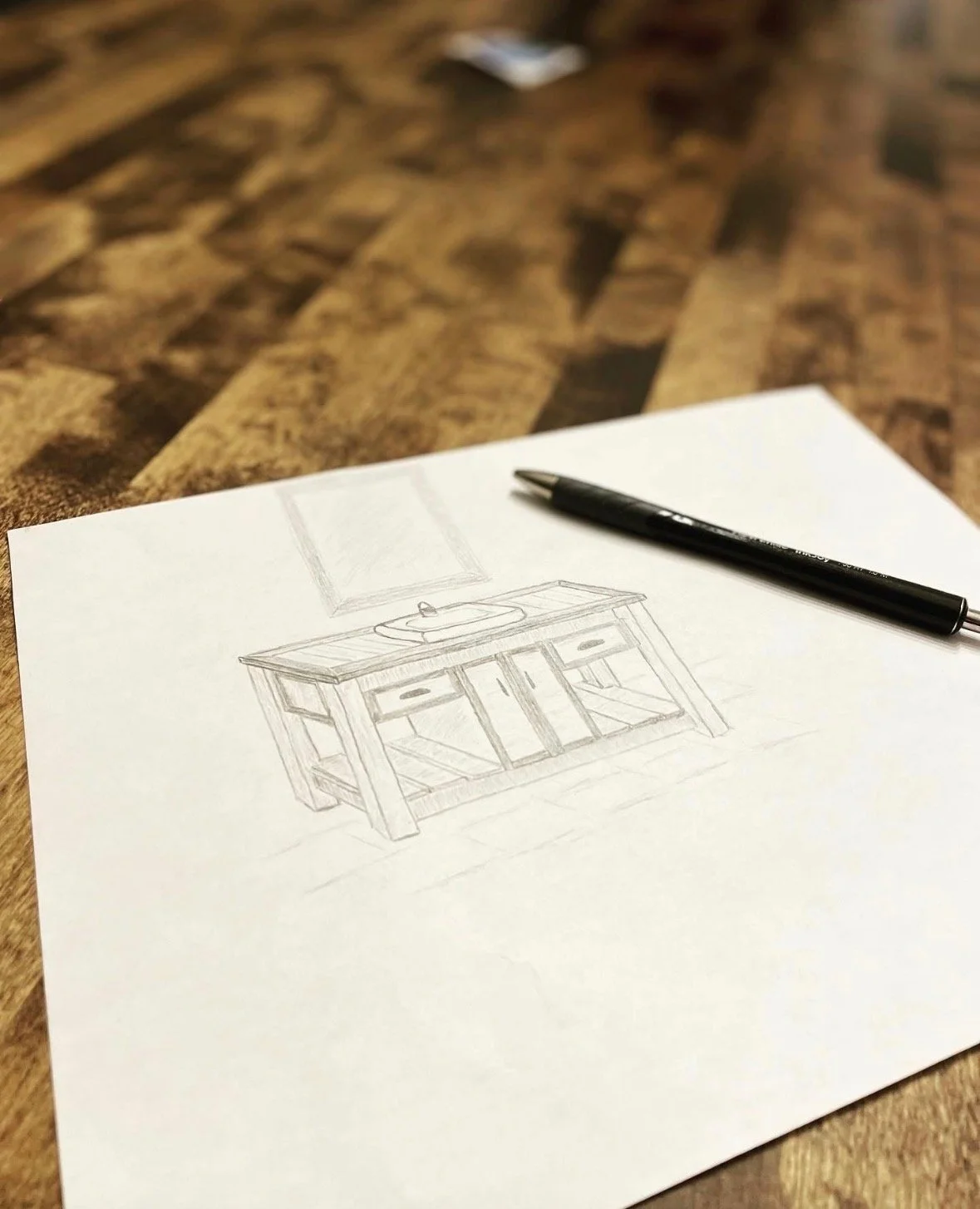 Sketch of a bathroom vanity with a sink and mirror on a piece of paper, with a black pen on a wooden table.
