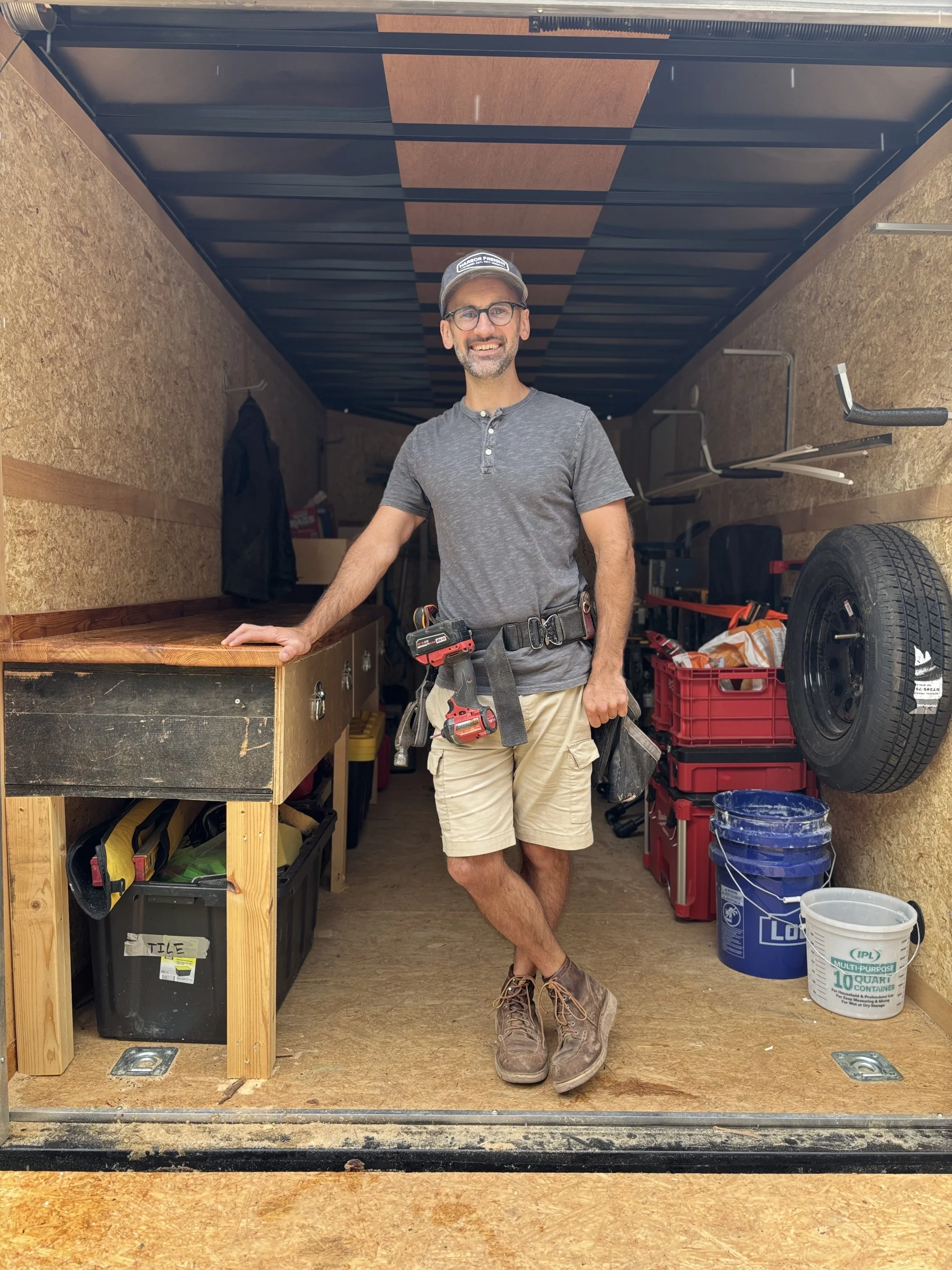 A man stands inside a storage trailer, smiling, dressed casually with glasses, a hat, and work boots. He has tools attached to his belt, and there are shelves, containers, a spare tire, and other work supplies around him.