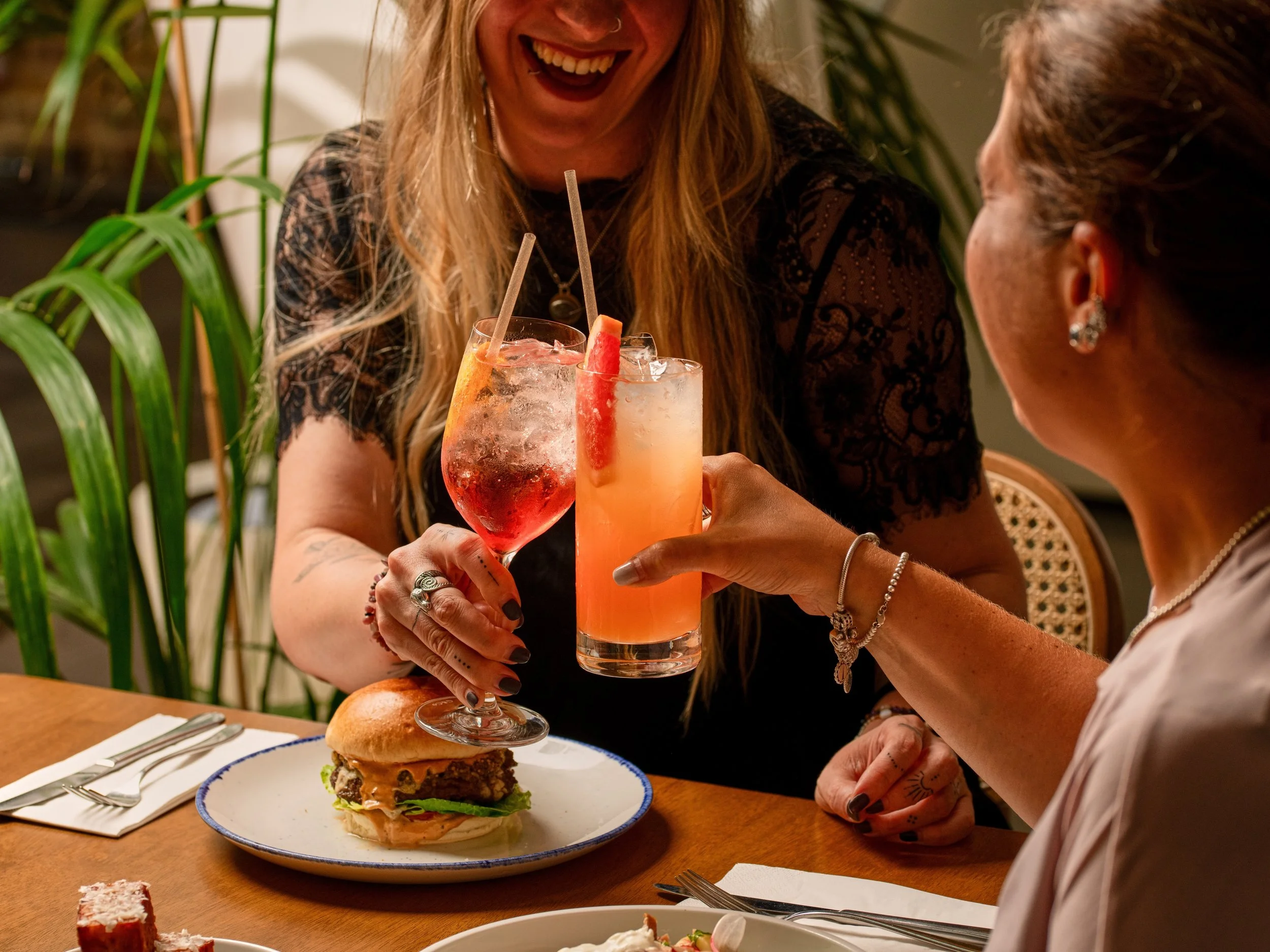 Two women sharing a toast with cocktails at a dinner table, with a burger on a plate in front of them, in a cozy indoor setting with plants.